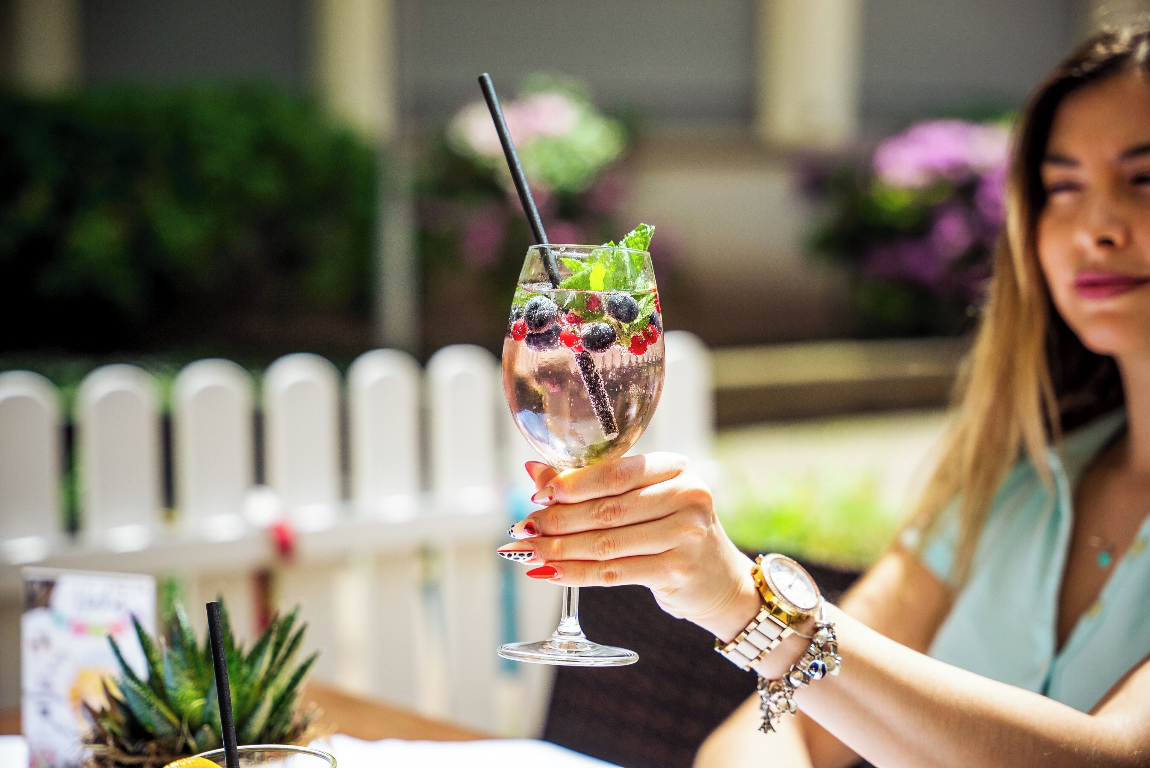 Une personne tenant un verre contenant une boisson décorée de baies et de menthe, assise à une table en plein air, avec une clôture blanche et de la verdure en arrière-plan.