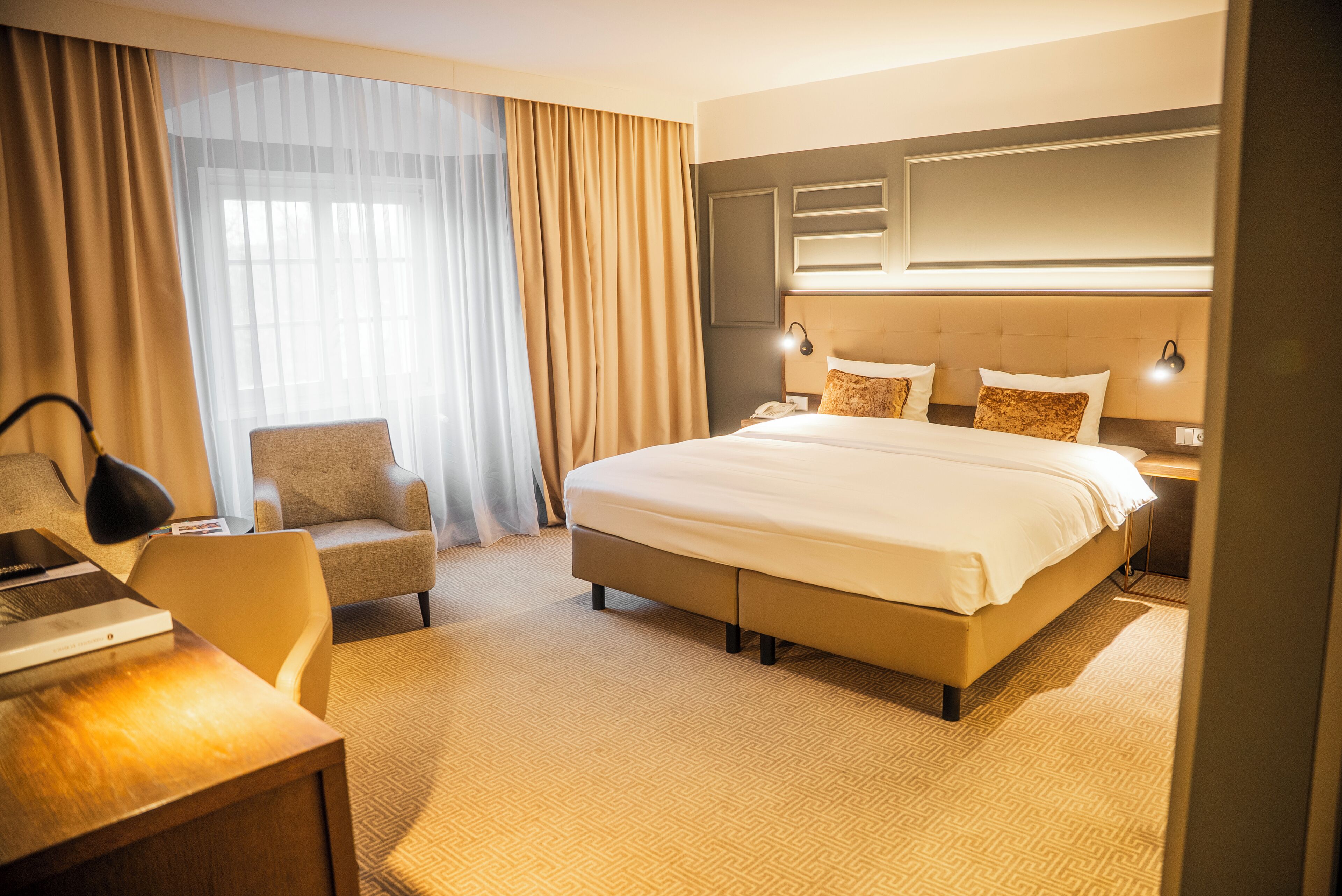 A hotel room with a large bed, a desk, a chair, and a nightstand with a lamp.