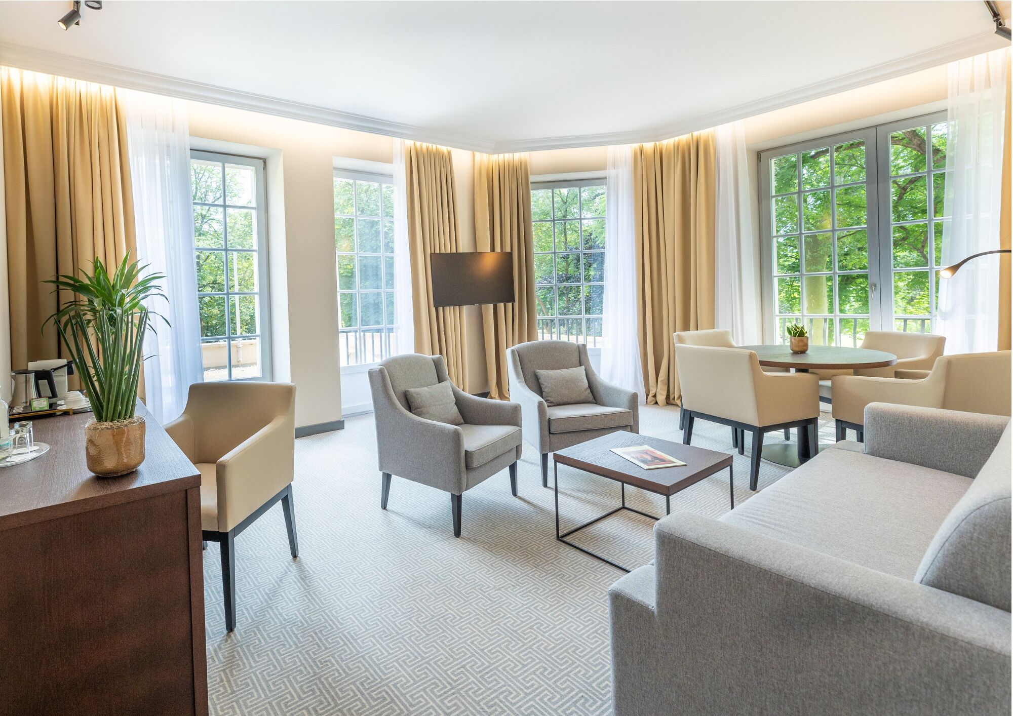 Ein helles, geräumiges Wohnzimmer mit Sofa, Sesseln, Couchtisch und Fernseher. Große Fenster mit Vorhängen lassen natürliches Licht und Blick auf die grüne Umgebung herein.