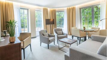 Ein helles, geräumiges Wohnzimmer mit Sofa, Sesseln, Couchtisch und Fernseher. Große Fenster mit Vorhängen lassen natürliches Licht und Blick auf die grüne Umgebung herein.
