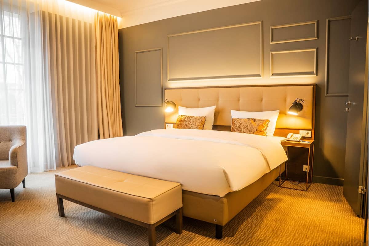 A hotel room with a large bed, a bedside table with a lamp, a chair, and a bench.