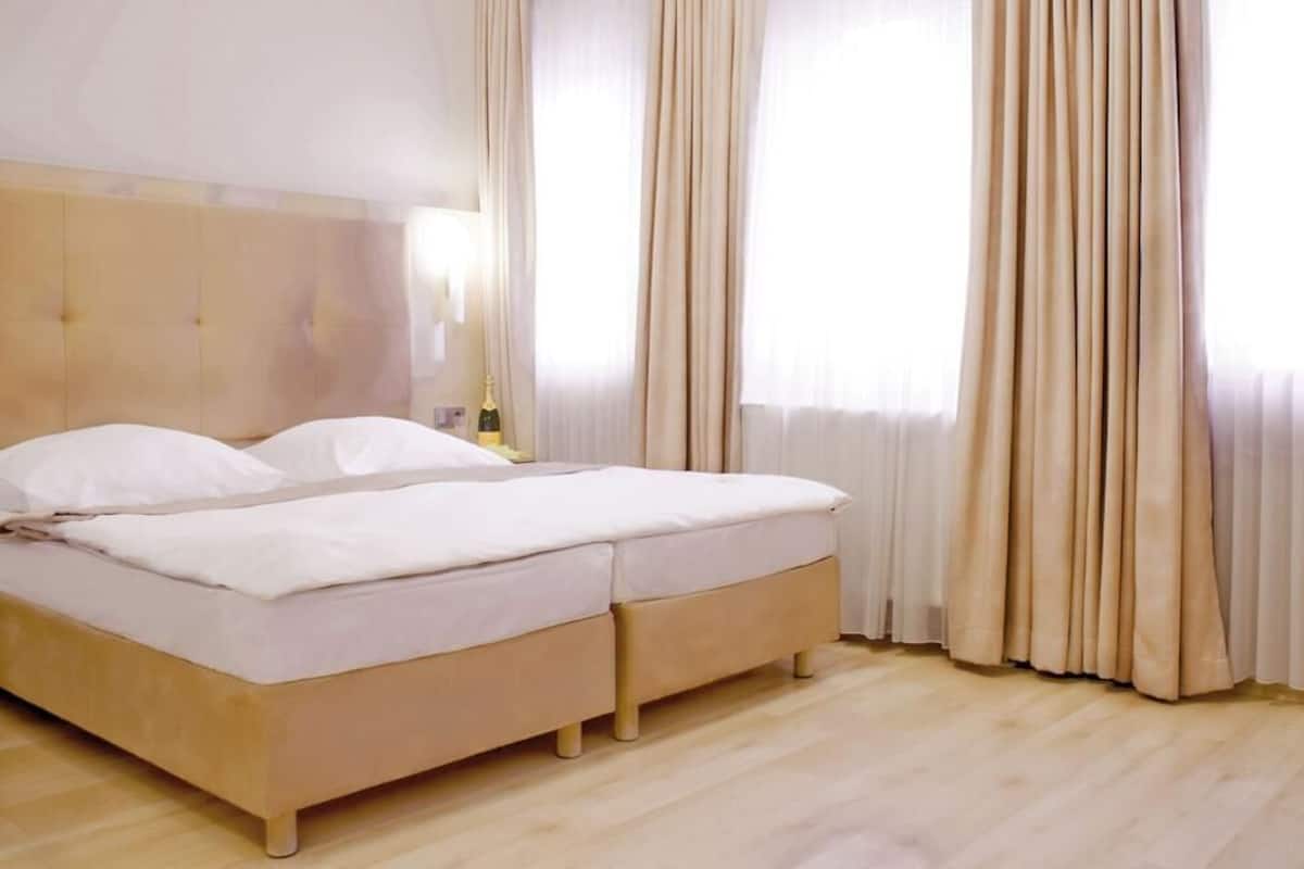 A double bed with a wooden headboard, a bedside table with a lamp, and sheer curtains.