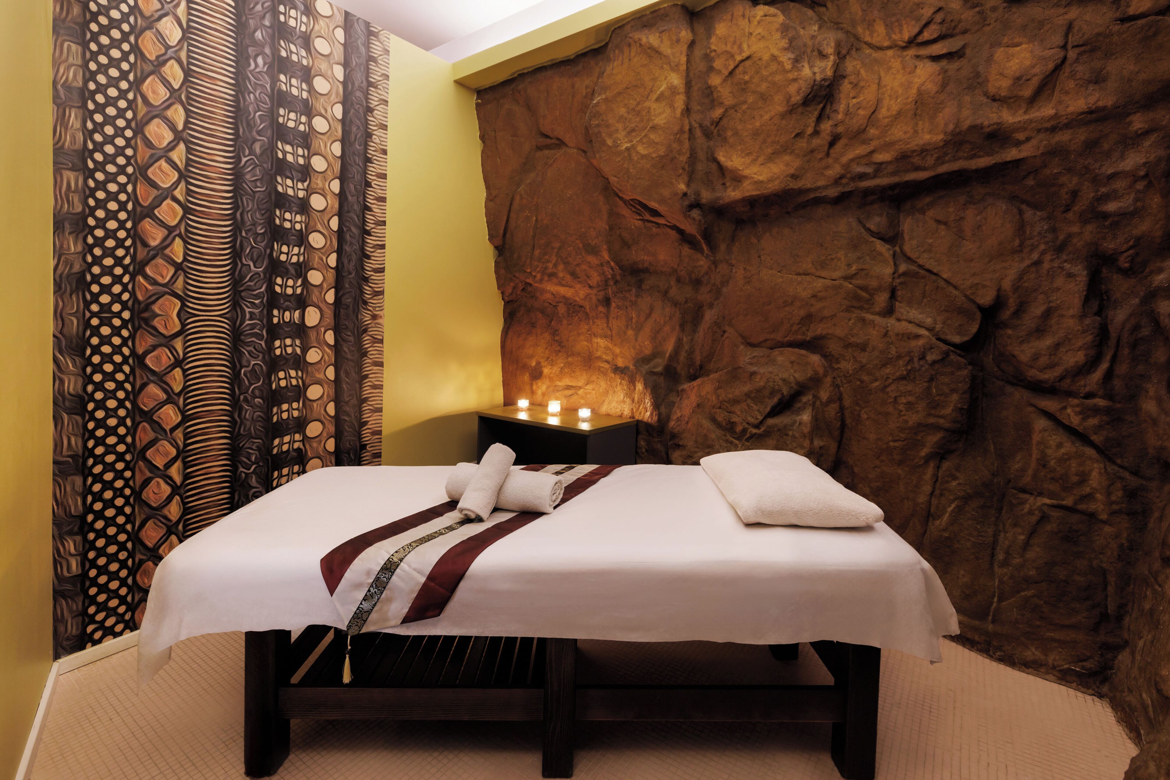A bed with a white bedspread and a red and white striped pillow, a nightstand with a lamp, and a wall with a rock-like texture.