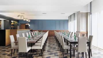 Un salón de conferencias con una larga mesa, sillas y una cocina al fondo.