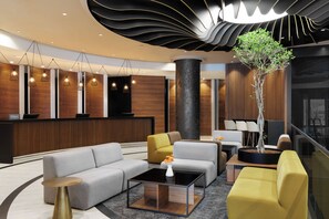 Een moderne hotel lobby met een receptiebalie, banken, een bijzettafeltje en een grote plantenbak.