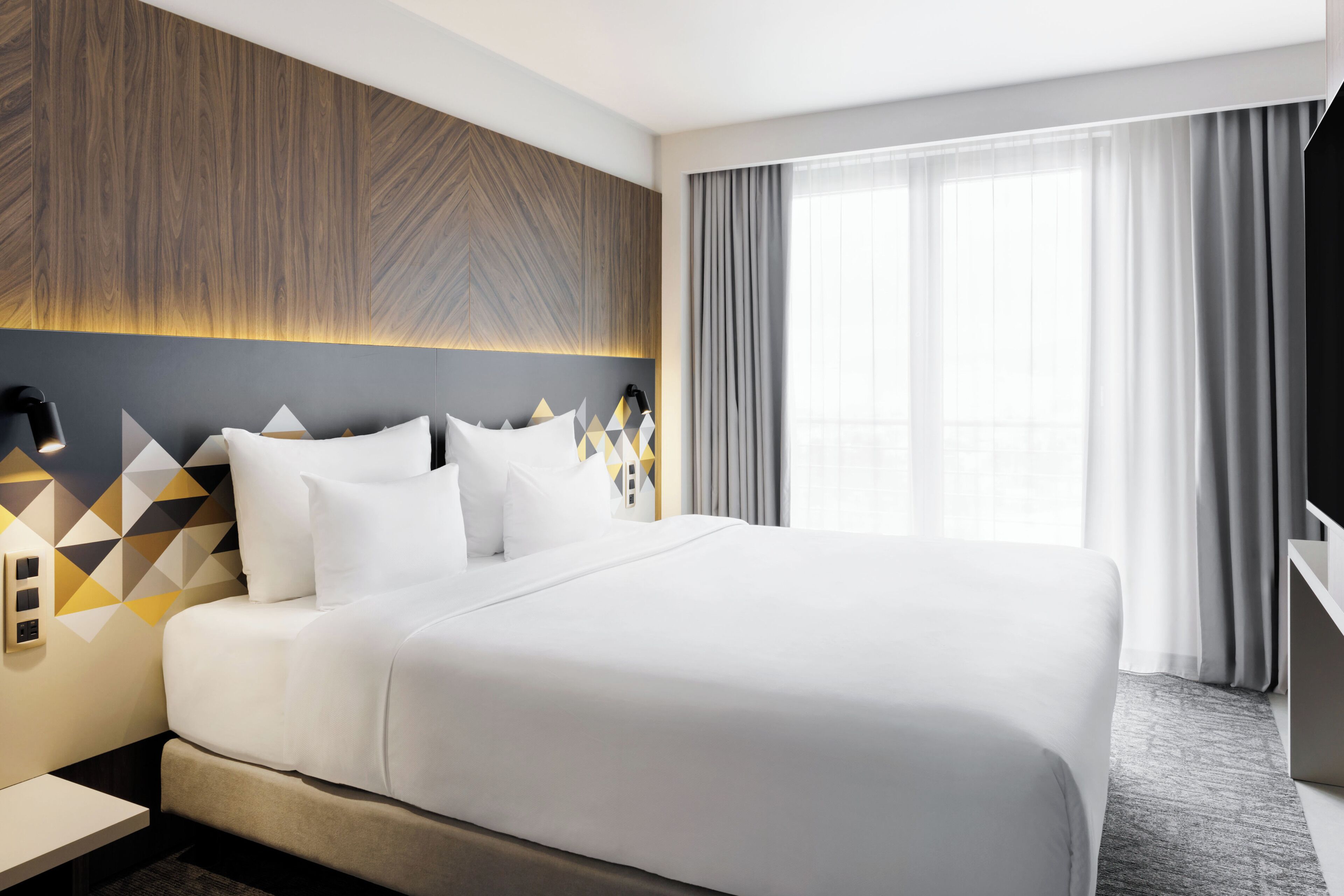 Ein modernes Hotelzimmer mit einem großen Bett, einem Kopfteil mit geometrischen Mustern, einem Nachttisch und Blick ins Freie durch ein Fenster mit Vorhängen.