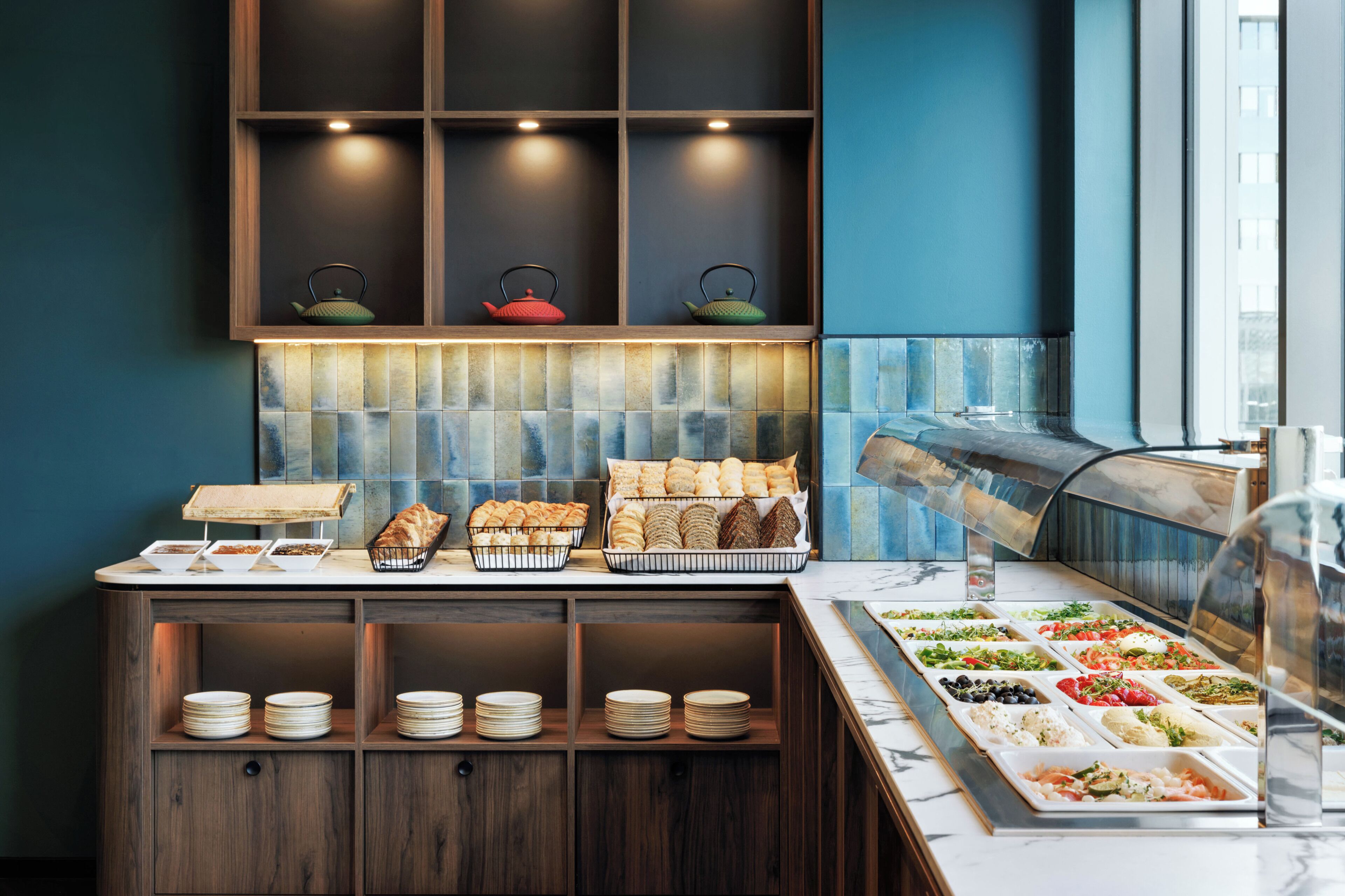 A buffet setup with various food items, plates, and baskets, with a window and a shelf with decorative items.