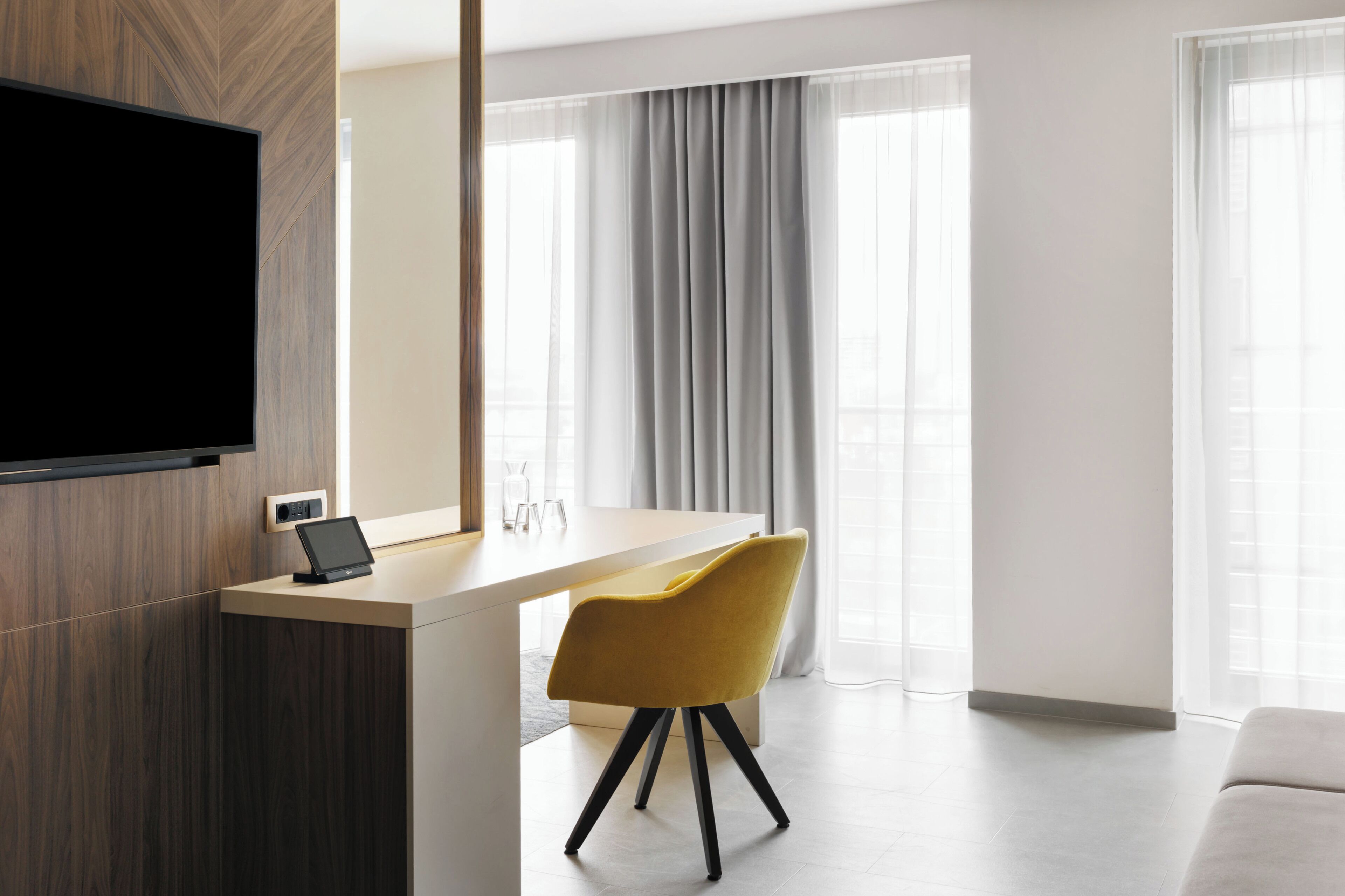 Ein modernes Hotelzimmer mit einem Flachbildfernseher, einem Schreibtisch mit einem Tablet, einem gelben Sessel und großen Fenstern mit Vorhängen.