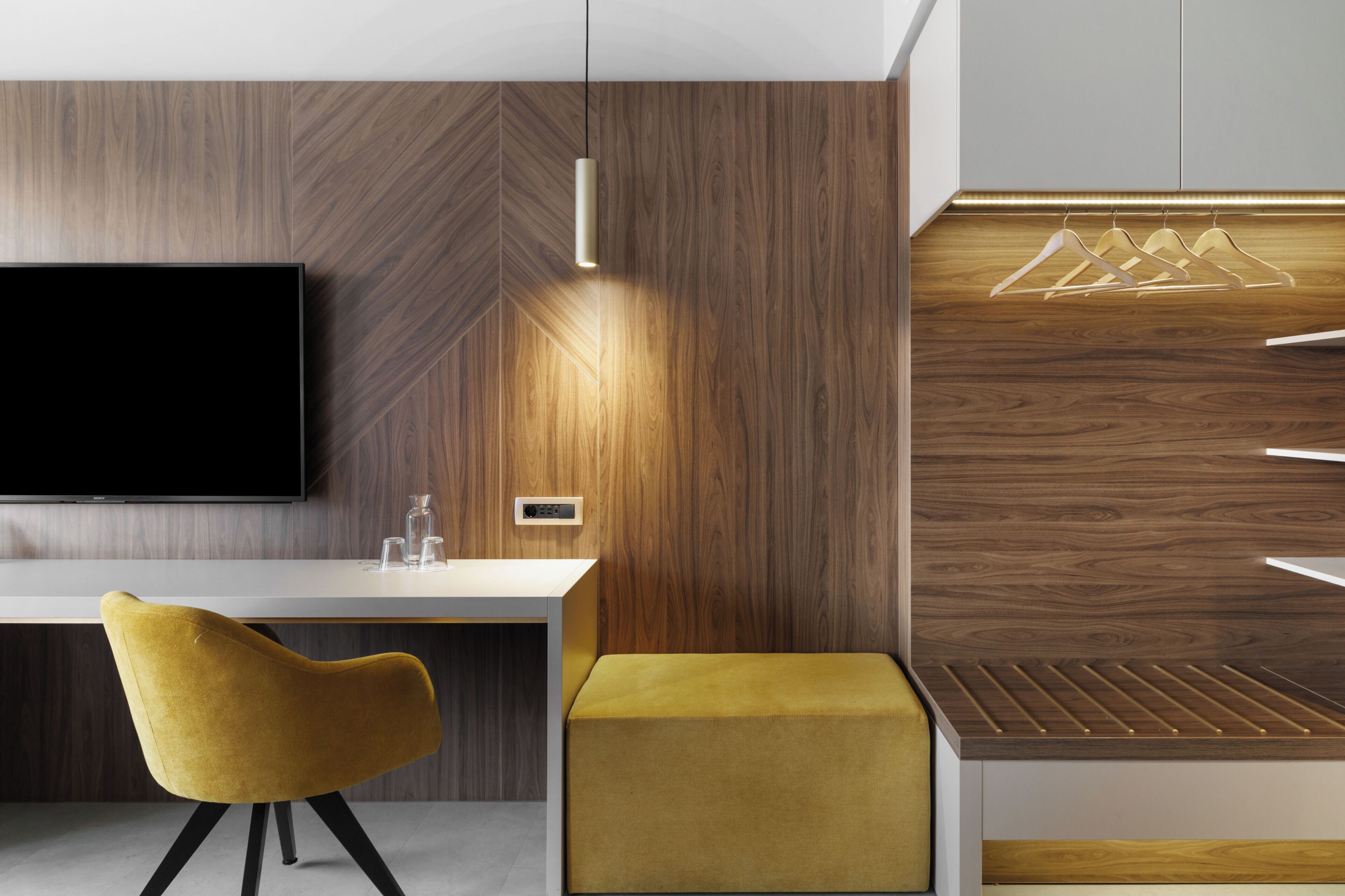A modern room with a flat-screen TV, a wooden accent wall, a yellow chair, a small ottoman, and a wooden shelf with hangers.
