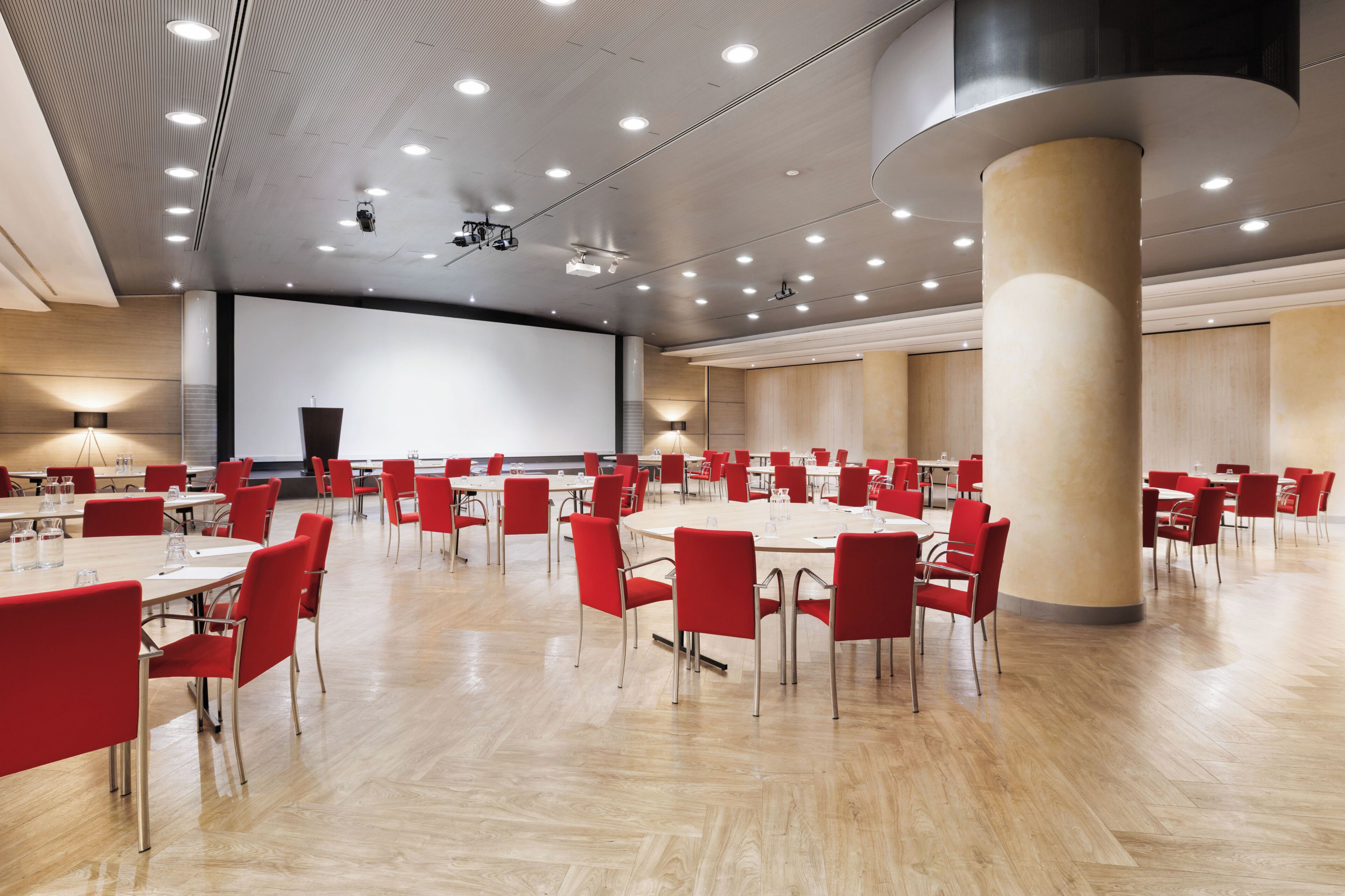 Una sala conferenze spaziosa con tavoli rotondi, sedie rosse e un grande schermo.