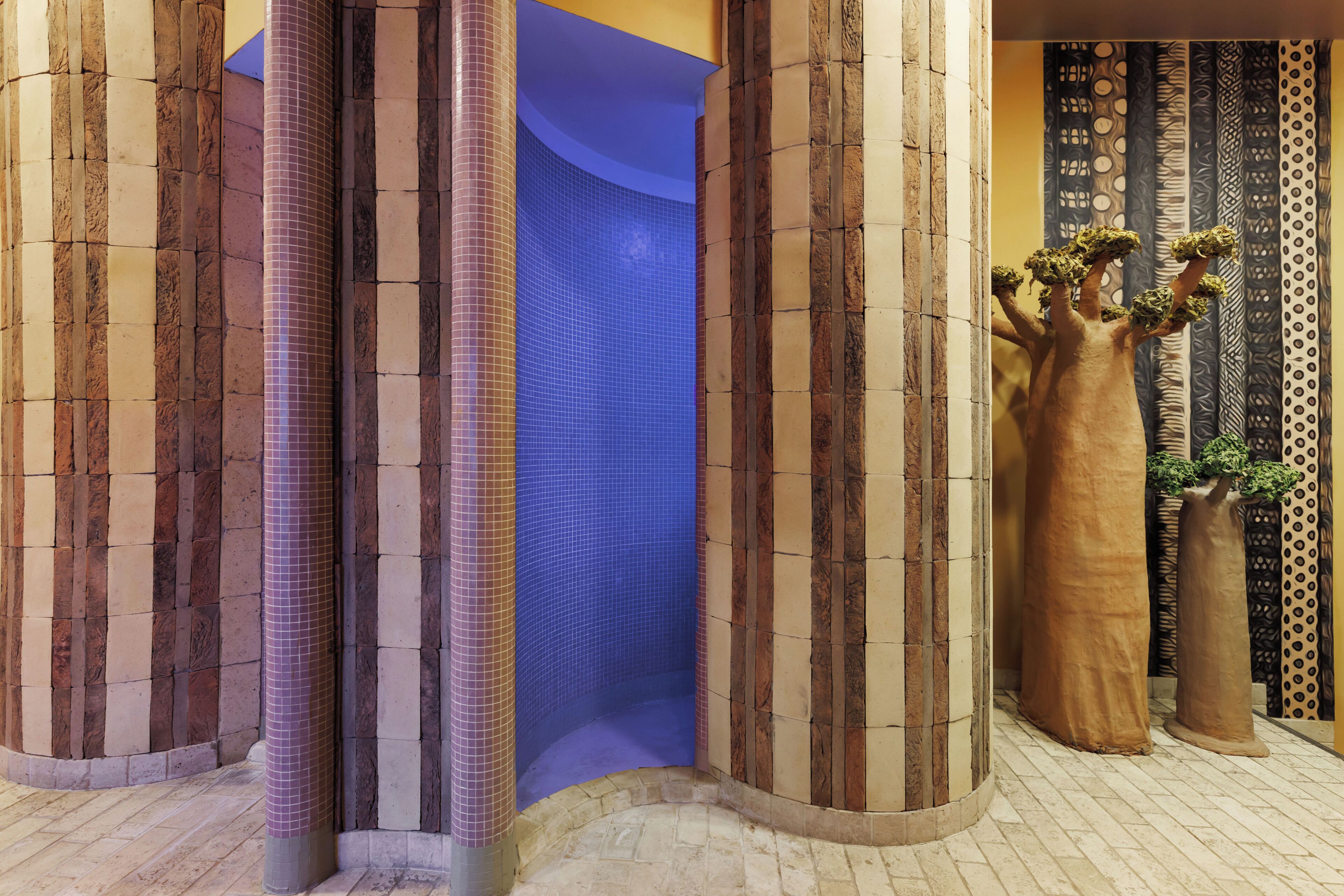 A corner with a blue-tiled wall, a column with a checkered pattern, and a large vase with a plant.