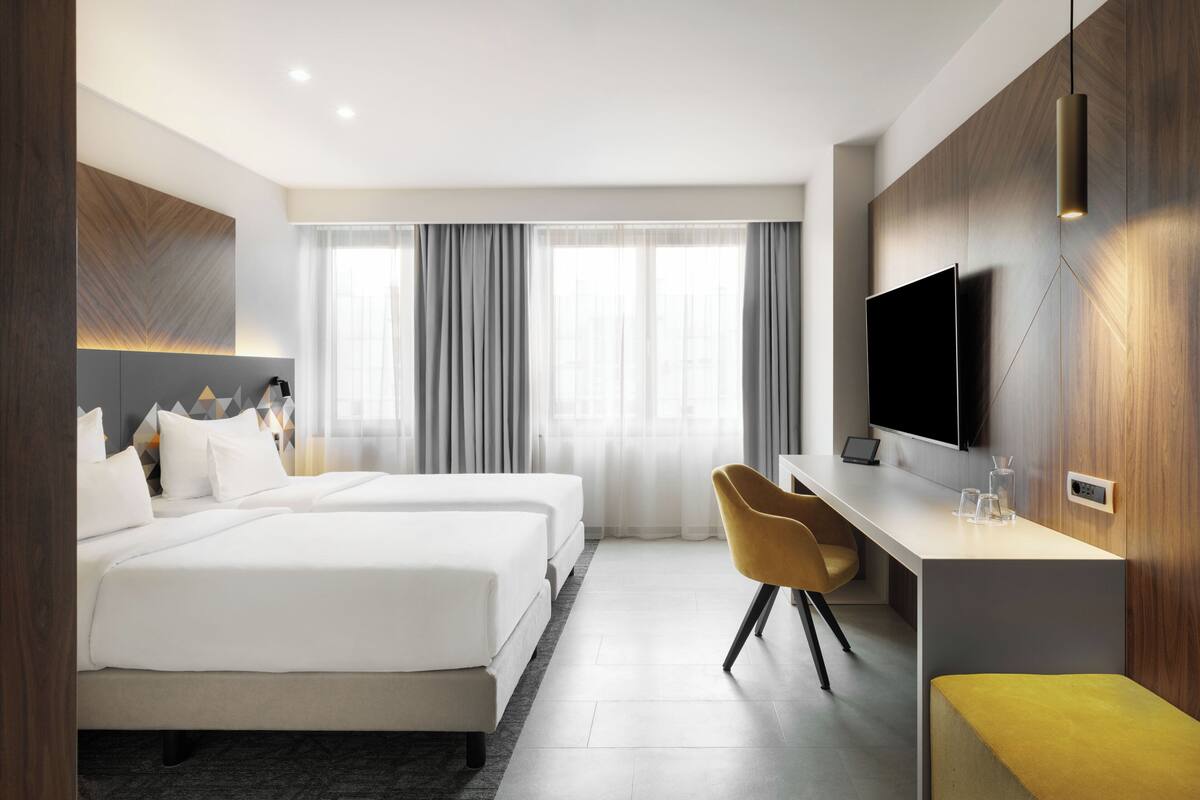 Ein Hotelzimmer mit zwei Betten, einem Schreibtisch, einem Stuhl, einem Fernseher und einem Fenster mit Vorhängen.