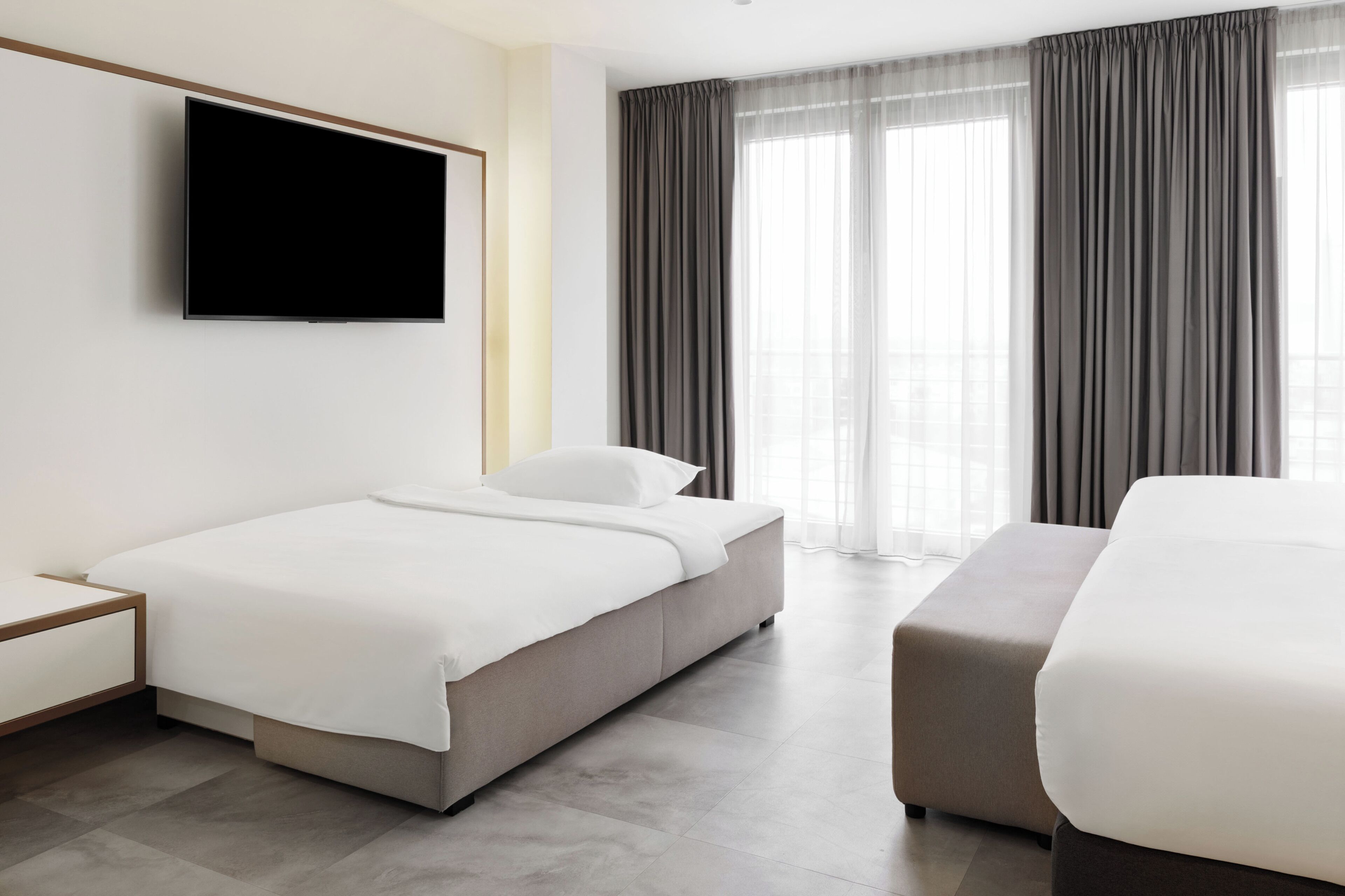 A modern hotel room with a bed, a bedside table, a flat-screen TV mounted on the wall, and a large window with curtains.