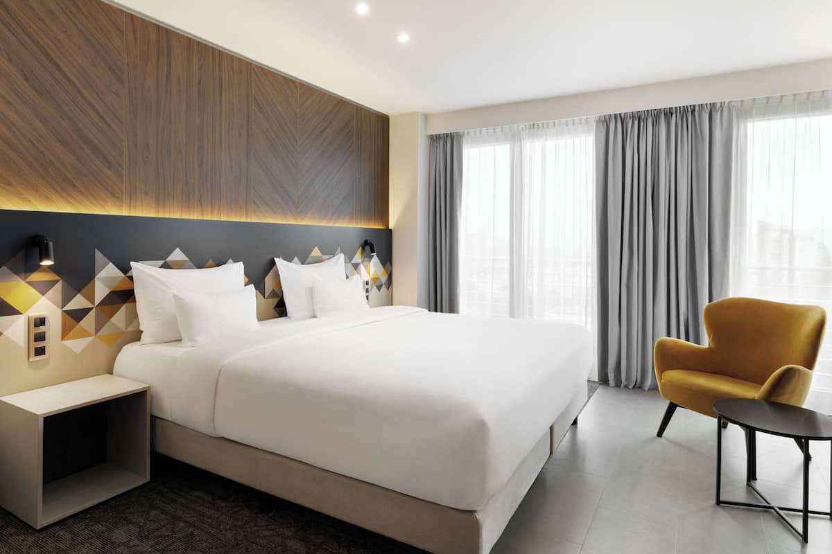 Ein modernes Hotelzimmer mit einem großen Bett, einem Nachttisch, einem Sessel und Blick auf die Stadt durch große Fenster.