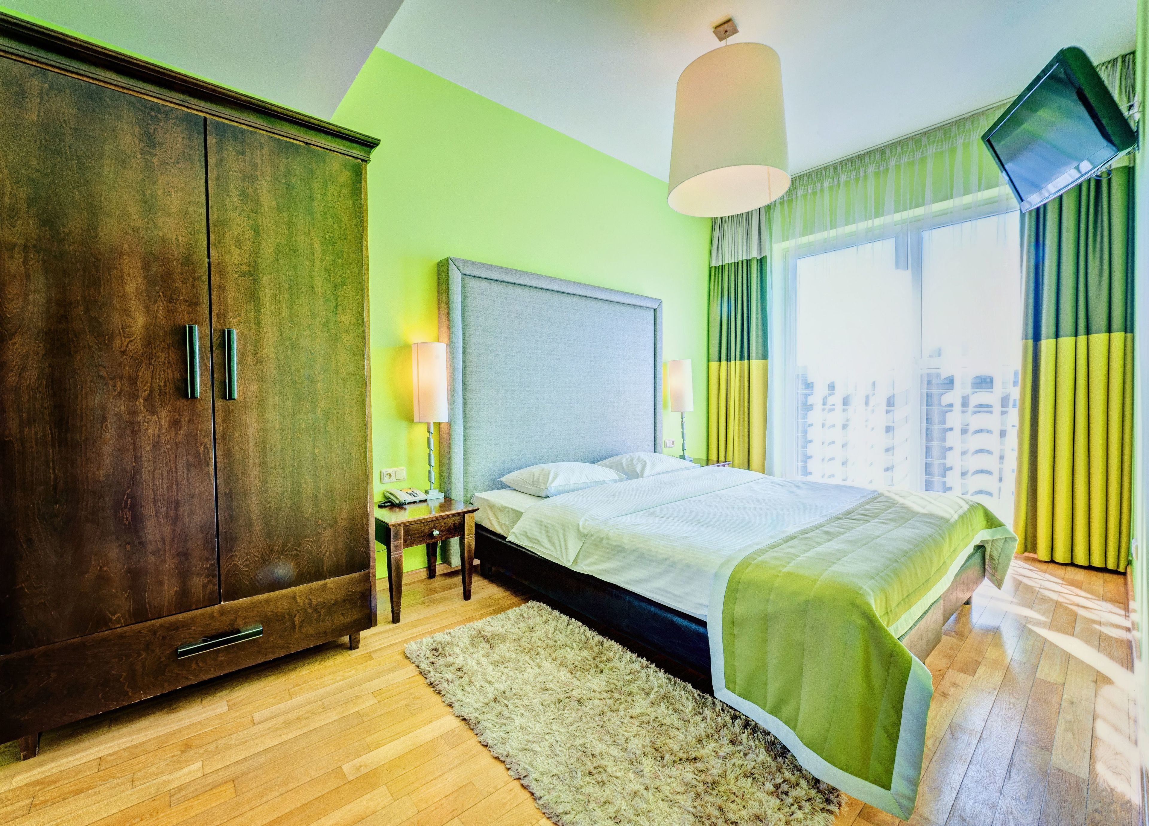 Une chambre d’hôtel dotée d’une grande armoire, d’un lit recouvert d’une couverture verte, d’une table de chevet avec une lampe et d’un téléviseur fixé au mur.