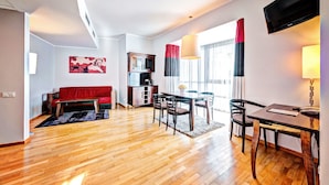 A modern living room with a wooden floor, a red sofa, a dining table with chairs, a TV mounted on the wall, and a bookshelf. - Don Prestige Residence (Poznan)