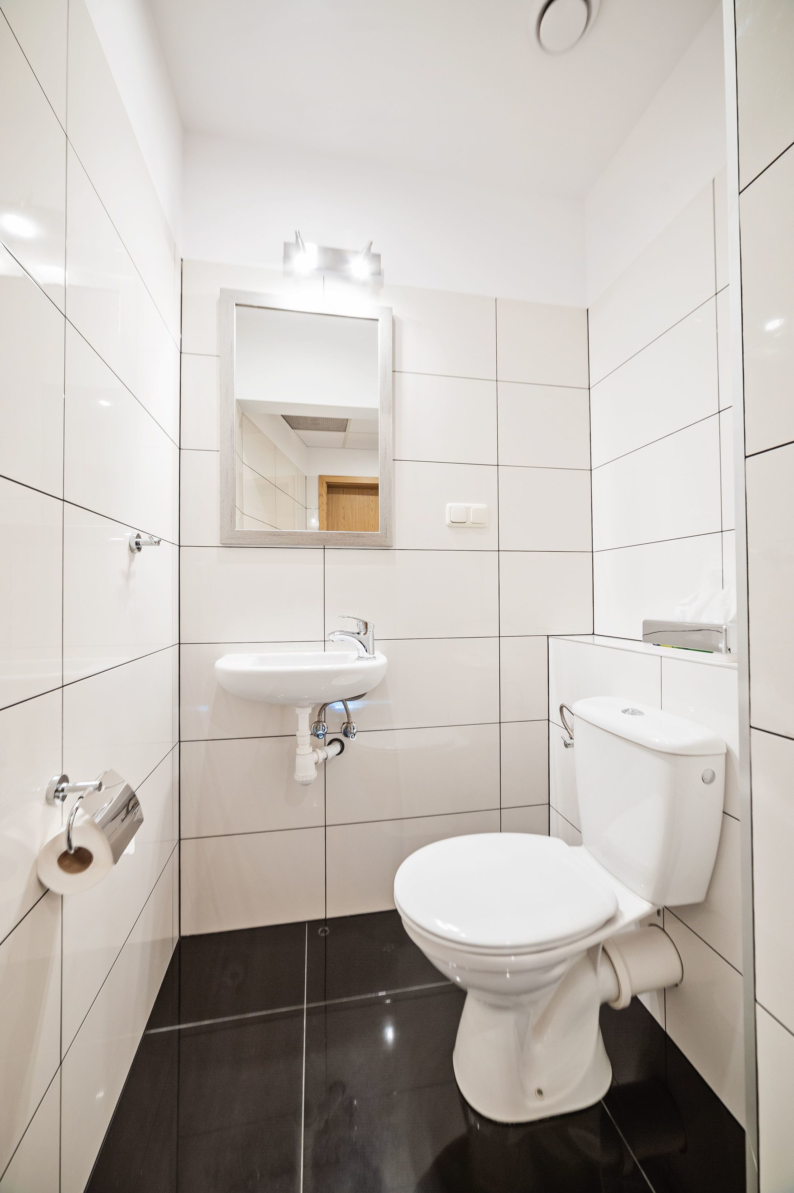 A modern bathroom with white tiles, a black floor, a white toilet, a sink, and a mirror.