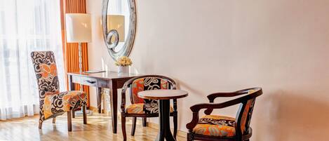 A hotel room with a wooden floor, a round table, two chairs with patterned upholstery, a large mirror, a television, and a lamp.