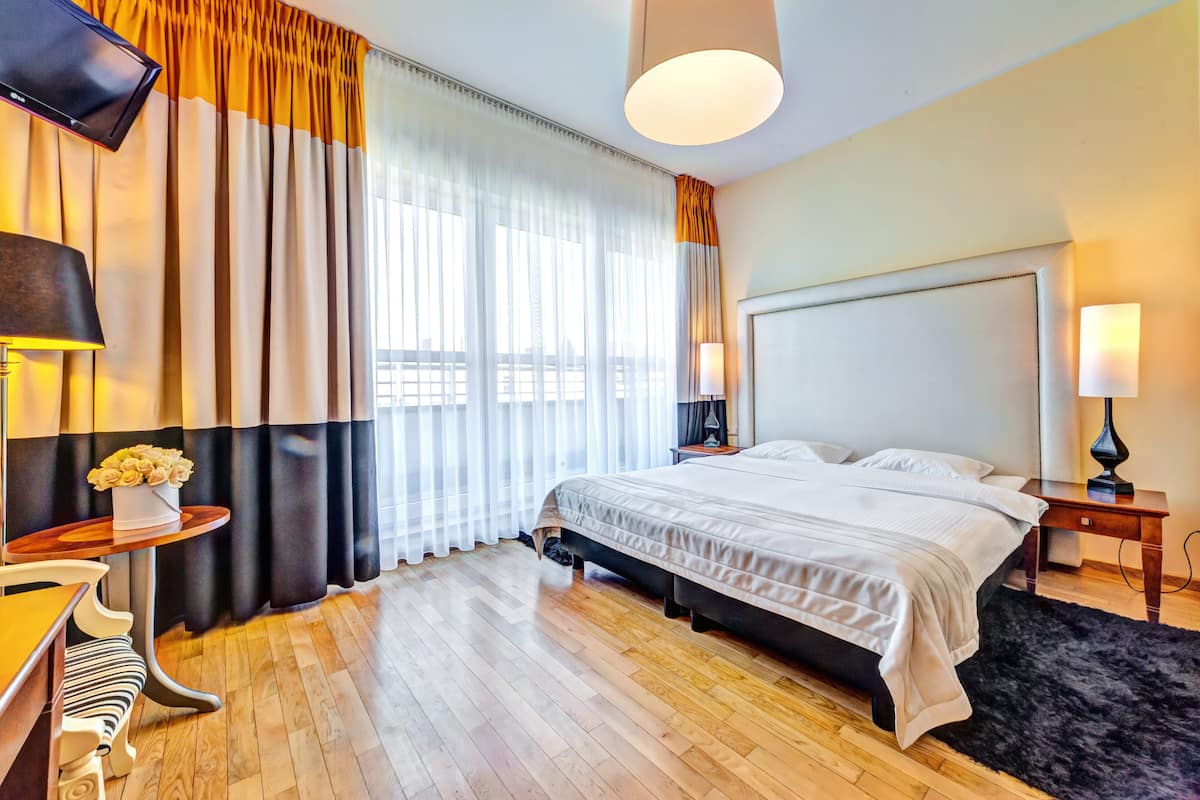 A hotel room with a large bed, a flat-screen TV mounted on the wall, a bedside table with a lamp, and a window with curtains.