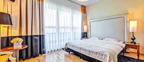 A hotel room with a large bed, a flat-screen TV mounted on the wall, a bedside table with a lamp, and a window with curtains.