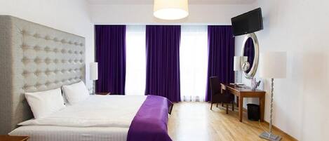 A modern hotel room with a large bed, a desk with a chair, a television, and a large window with purple curtains.
