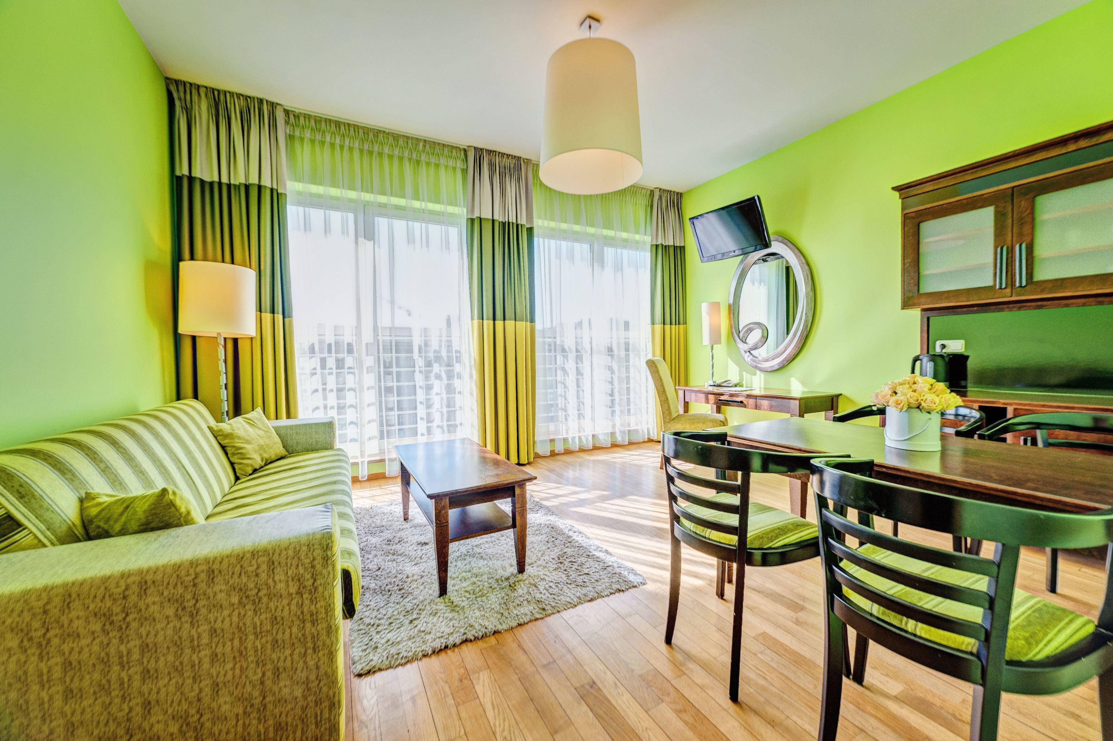 Ein modernes Hotelzimmer mit grüner Farbgestaltung, einer Sitzecke, einem Esstisch mit Stühlen, einem flachbildfernseher an der Wand und einem großen Fenster mit durchsichtigen Vorhängen.