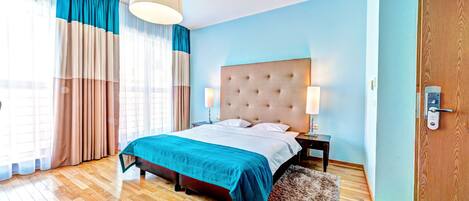 A bedroom with a bed, wooden flooring, a large window with curtains, a nightstand, and a door with a lock.
