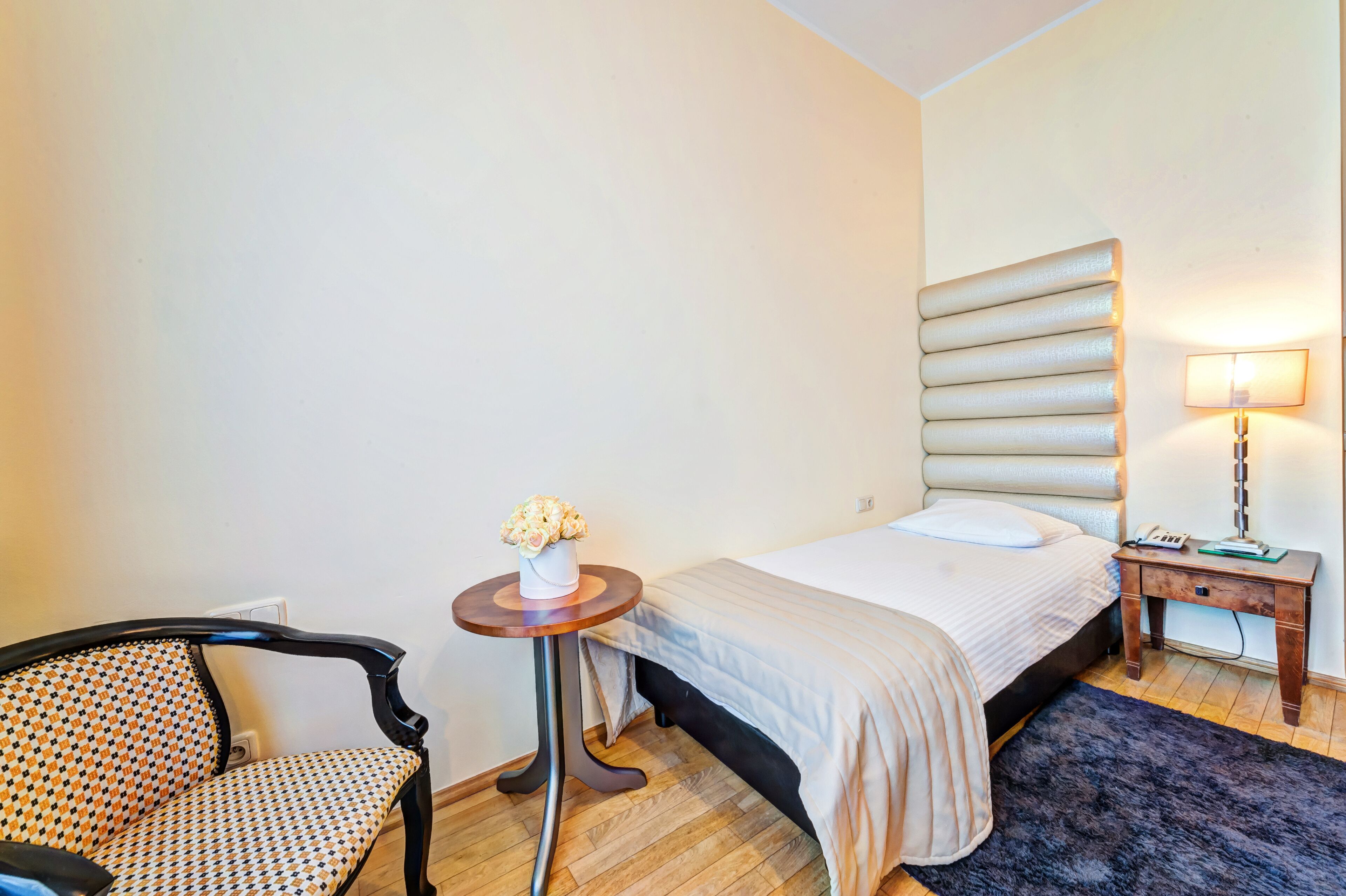 A hotel room with a bed, a chair, a small table with a vase of flowers, a nightstand with a lamp, and a wooden floor.