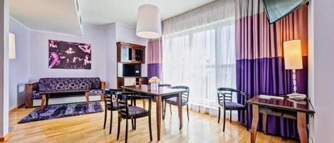 A modern living room with a sofa, dining table, chairs, a TV, and a large window with curtains.