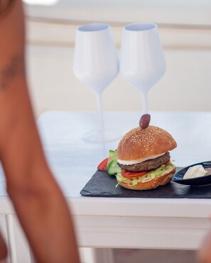 A burger with lettuce, tomato, and a sesame seed bun on a slate board, accompanied by two white wine glasses. - Domina Aquamarine Resort (Sharm El Sheikh)