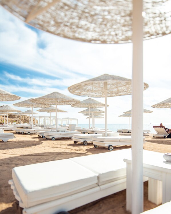 A beachside lounge area with white lounge chairs and umbrellas, a sandy beach, and a clear view of the sea.