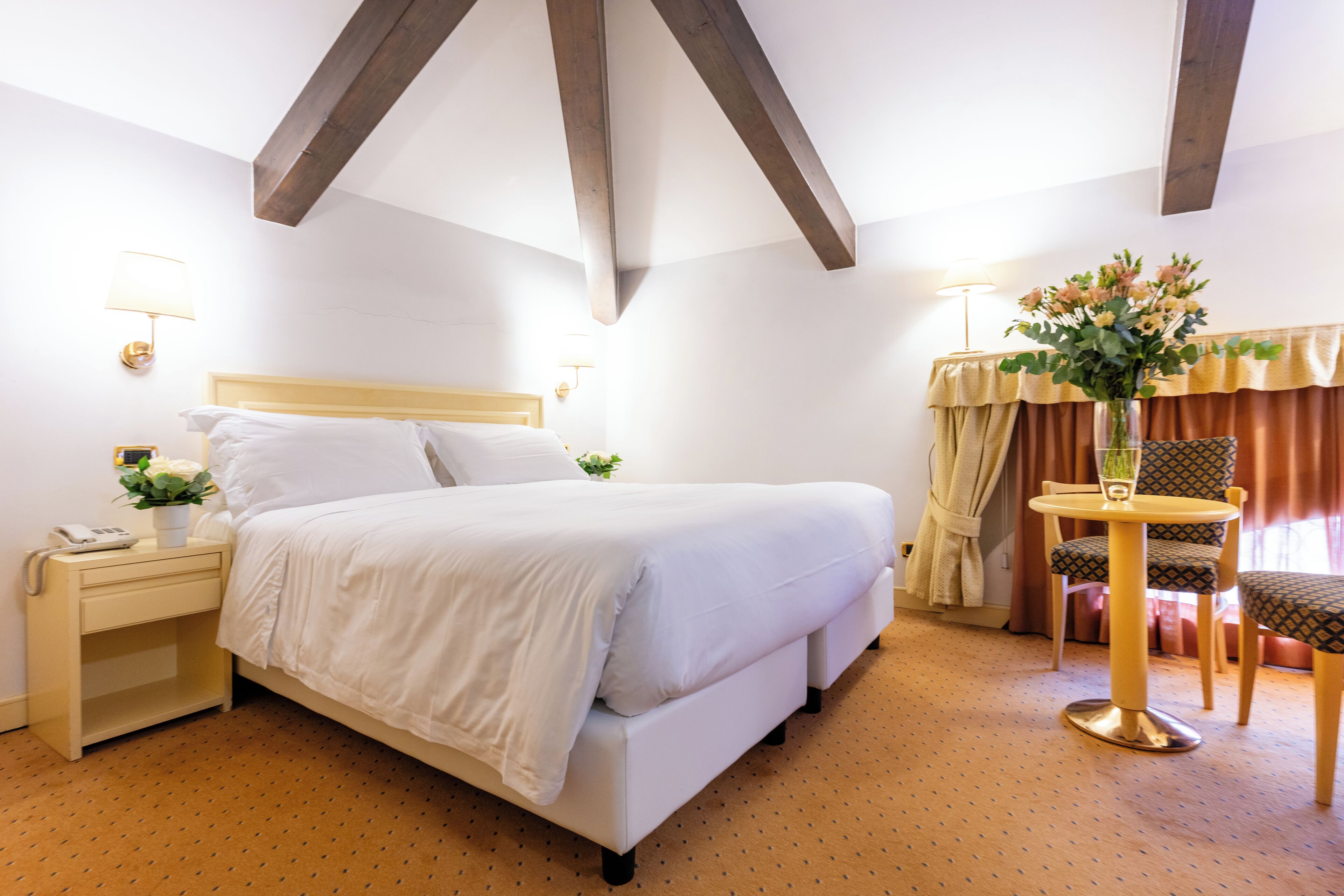 A hotel room with a bed, bedside table, lamp, chair, and a small table with a vase.