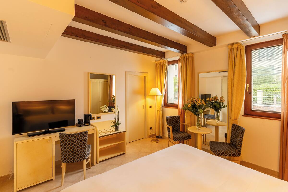 A hotel room with a bed, a desk with a TV, a chair, a small table with a lamp, and a window with curtains.