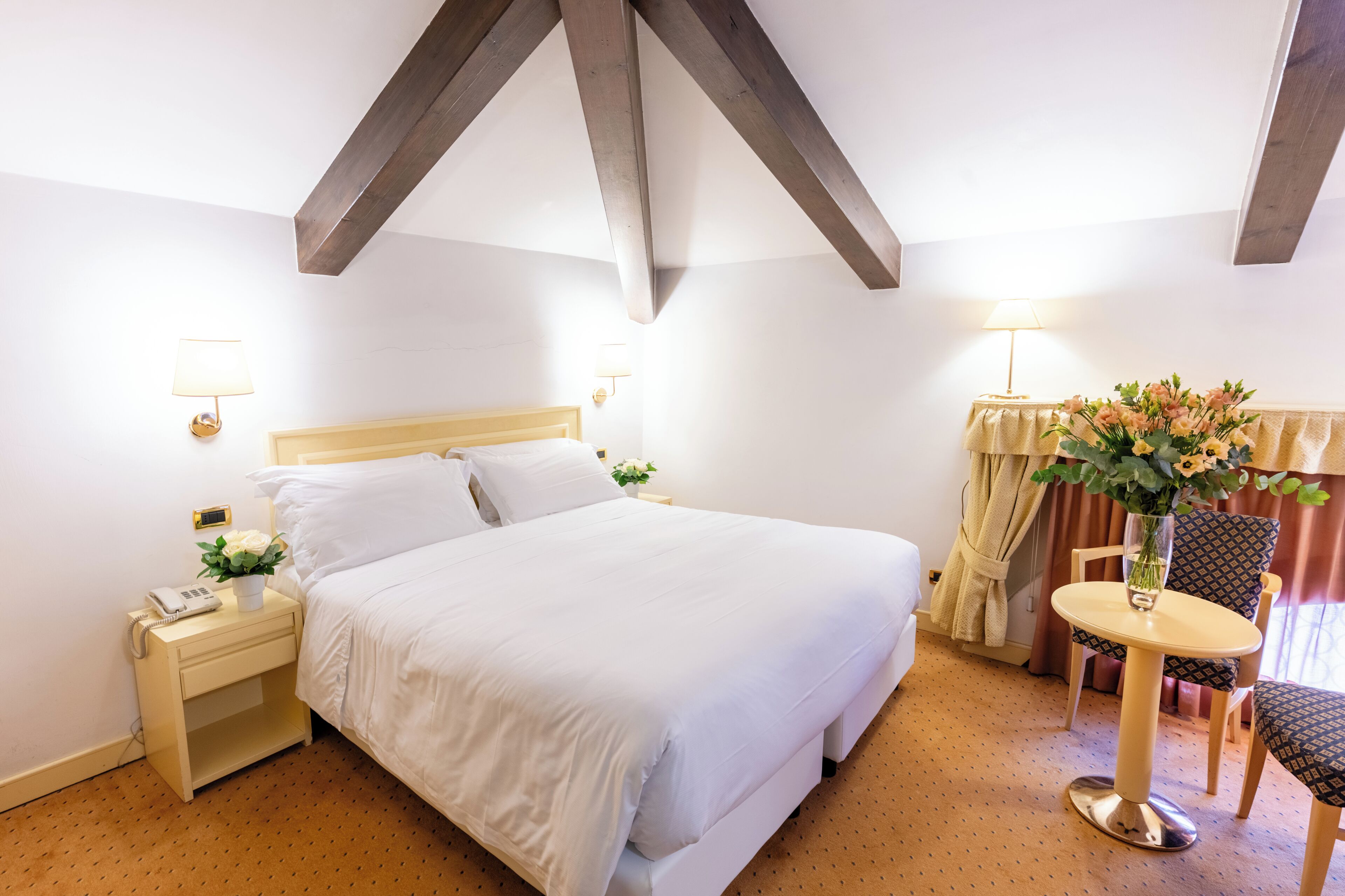 A hotel room with a bed, bedside table, lamp, chair, and a small table with a vase of flowers.