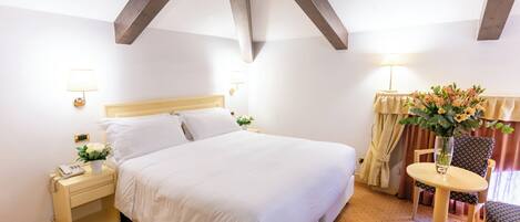 A hotel room with a bed, bedside table, lamp, chair, and a small table with a vase of flowers.