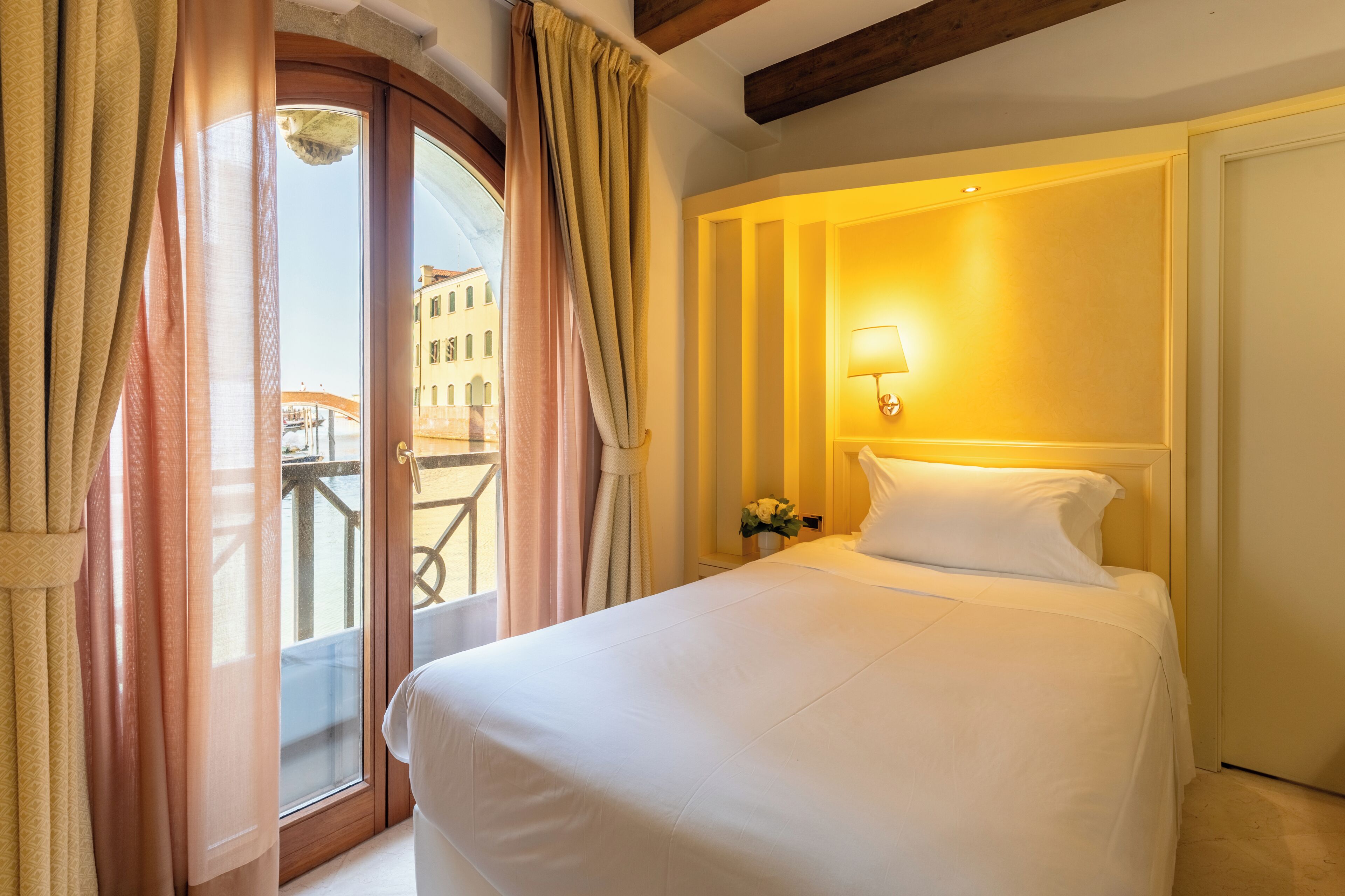 Ein Hotelzimmer mit Bett, einem Vorhangfenster, einer Wandlampe und Blick auf Gebäude.