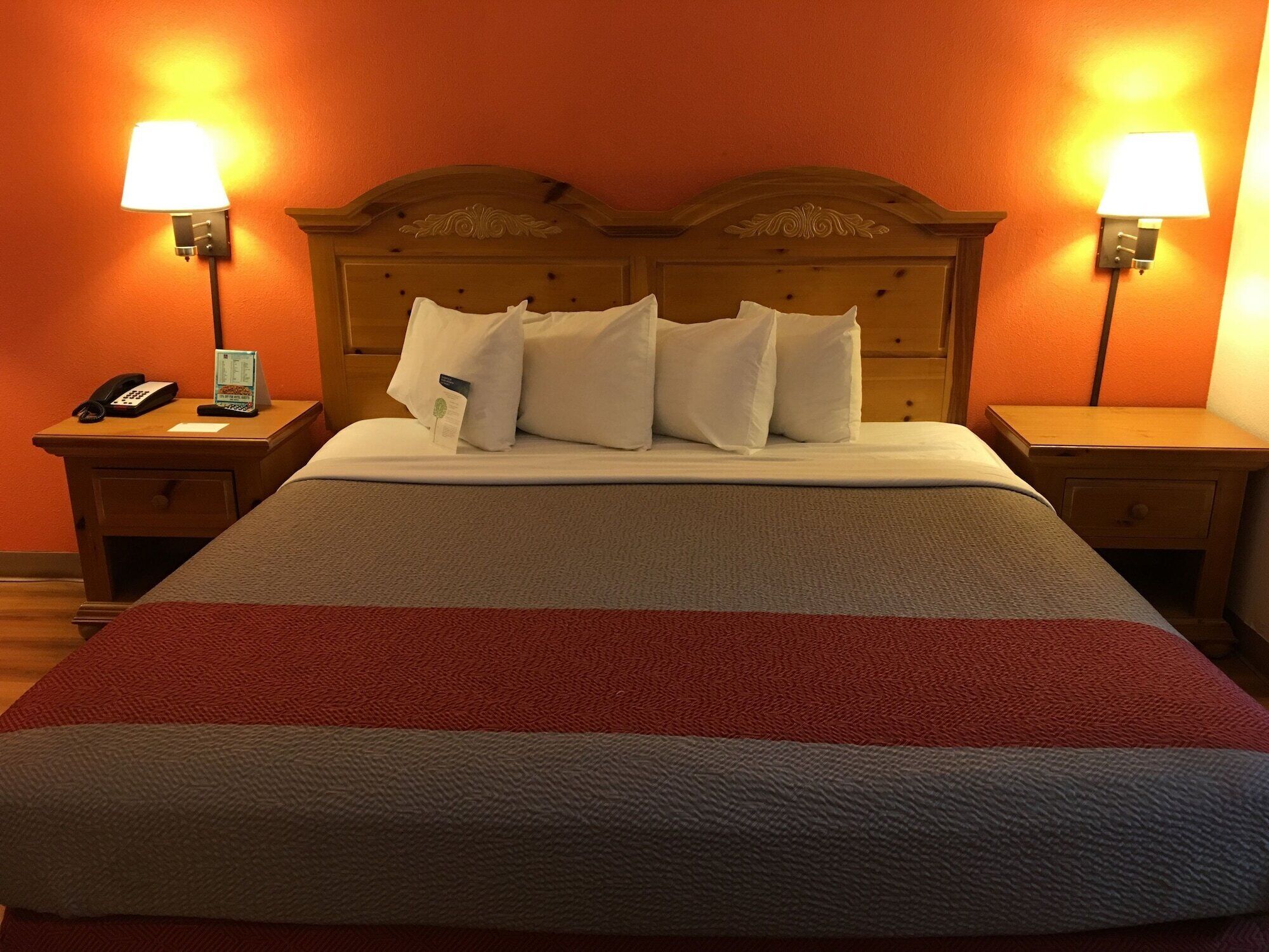 A hotel room with a wooden headboard, two bedside lamps, a telephone, and a neatly made bed with a red and gray blanket.