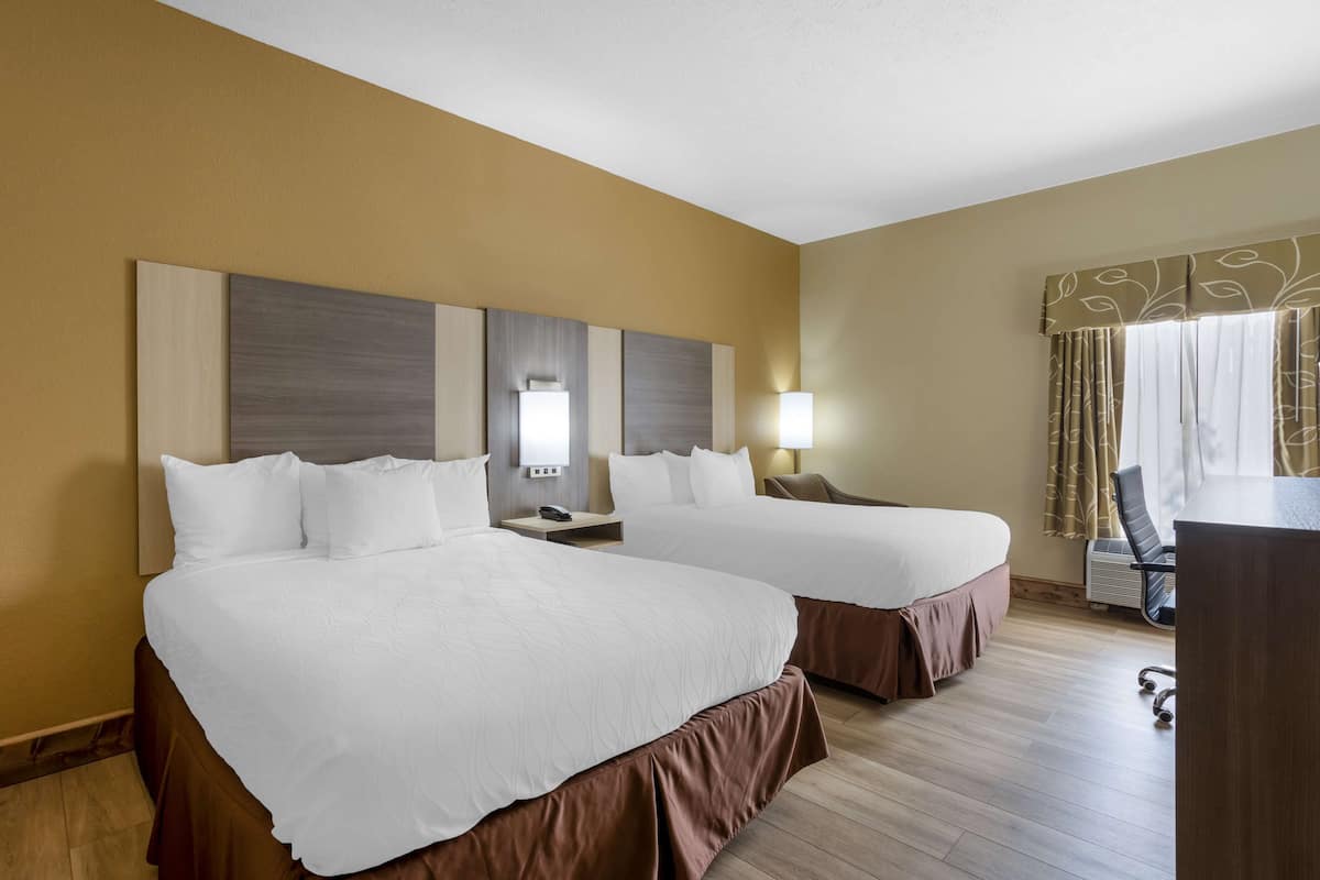 A hotel room with two beds, a desk, a chair, and a lamp.