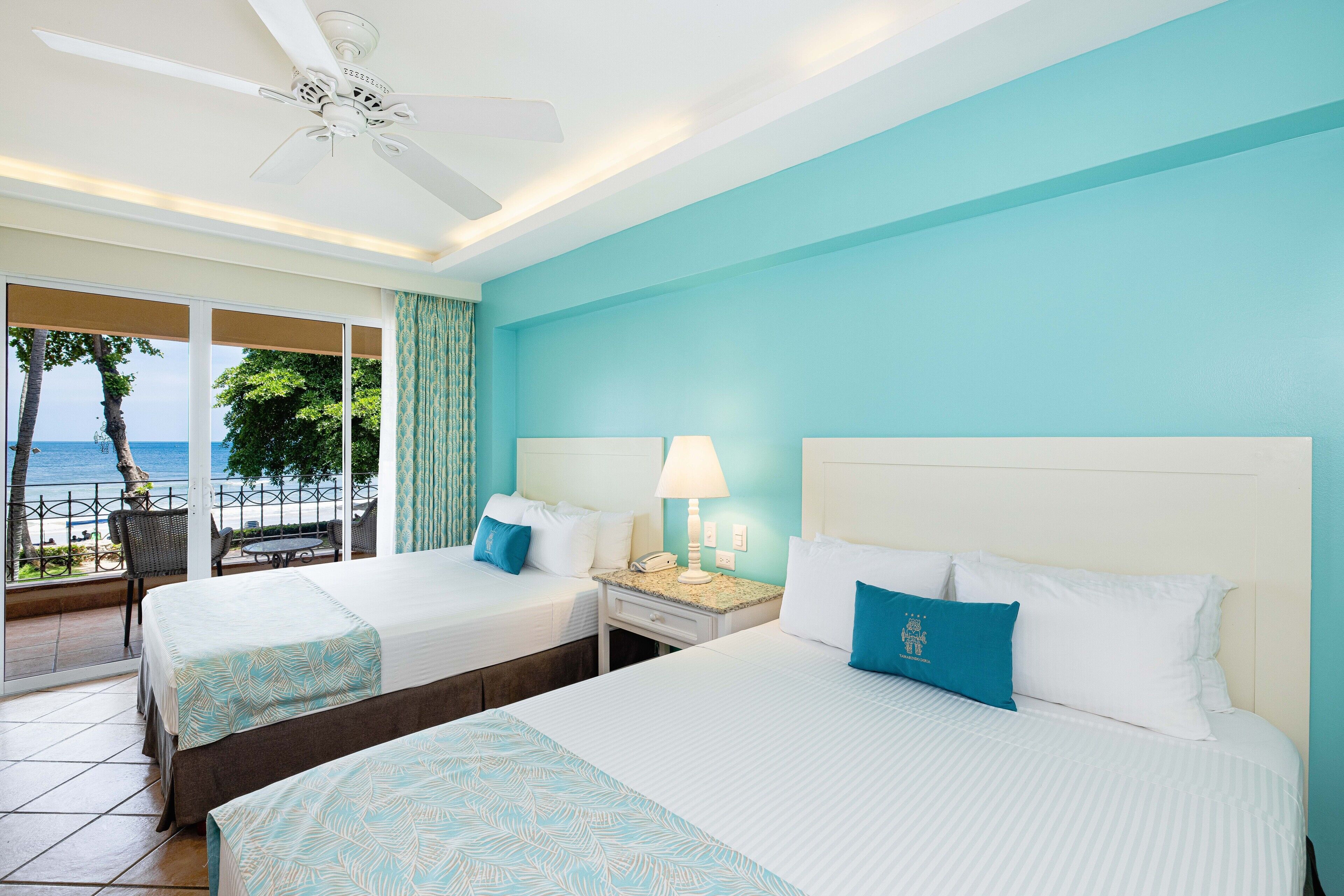 A hotel room with two beds, a ceiling fan, a nightstand with a lamp, and a view of the ocean.