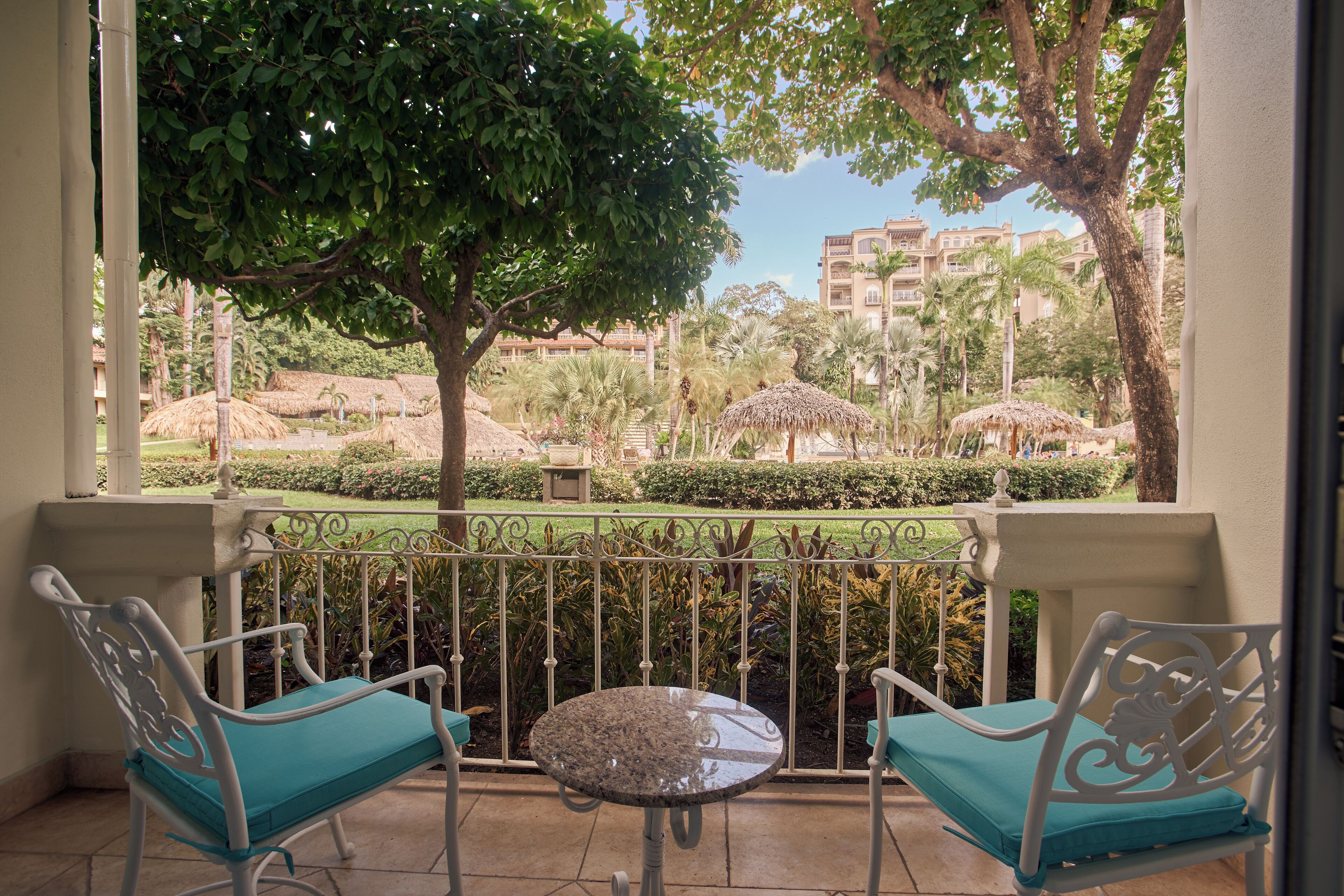 Ein Balkon mit zwei Stühlen und einem kleinen Tisch, der einen angelegten Garten mit Bäumen und einem Gebäude im Hintergrund zeigt.
