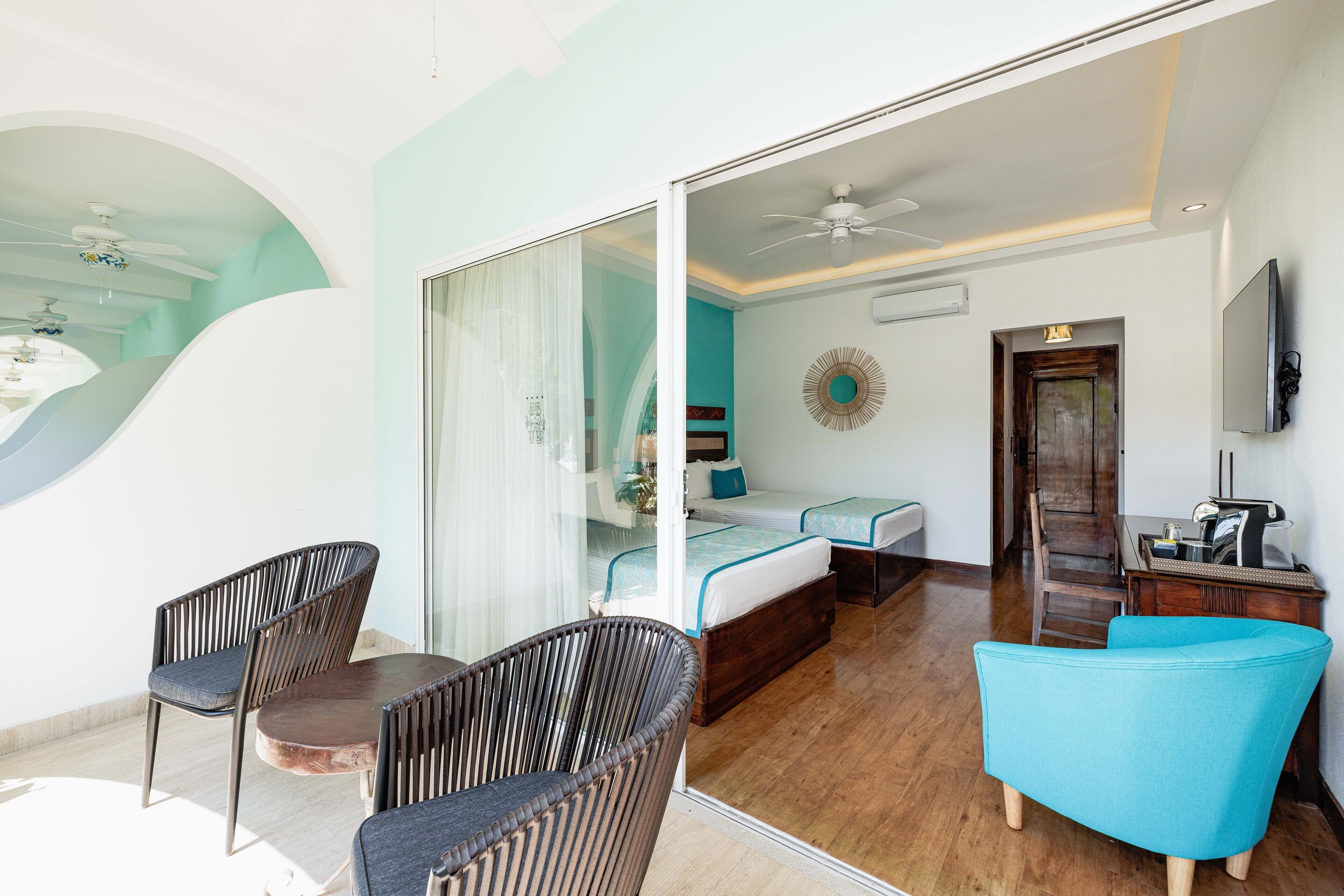 Una habitación de hotel con puerta de vidrio que da a un balcón, cama con detalles en azul, televisor y escritorio de madera.