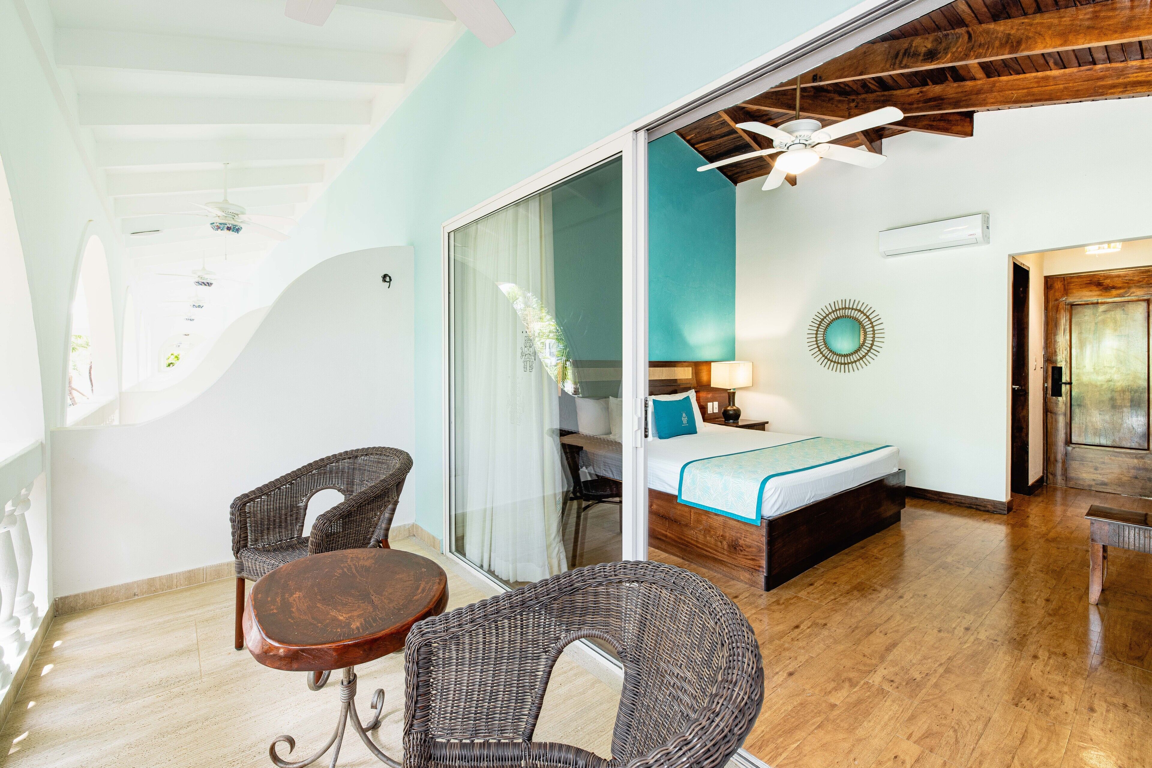 A room with a bed, a table, two wicker chairs, a ceiling fan, and a glass partition.