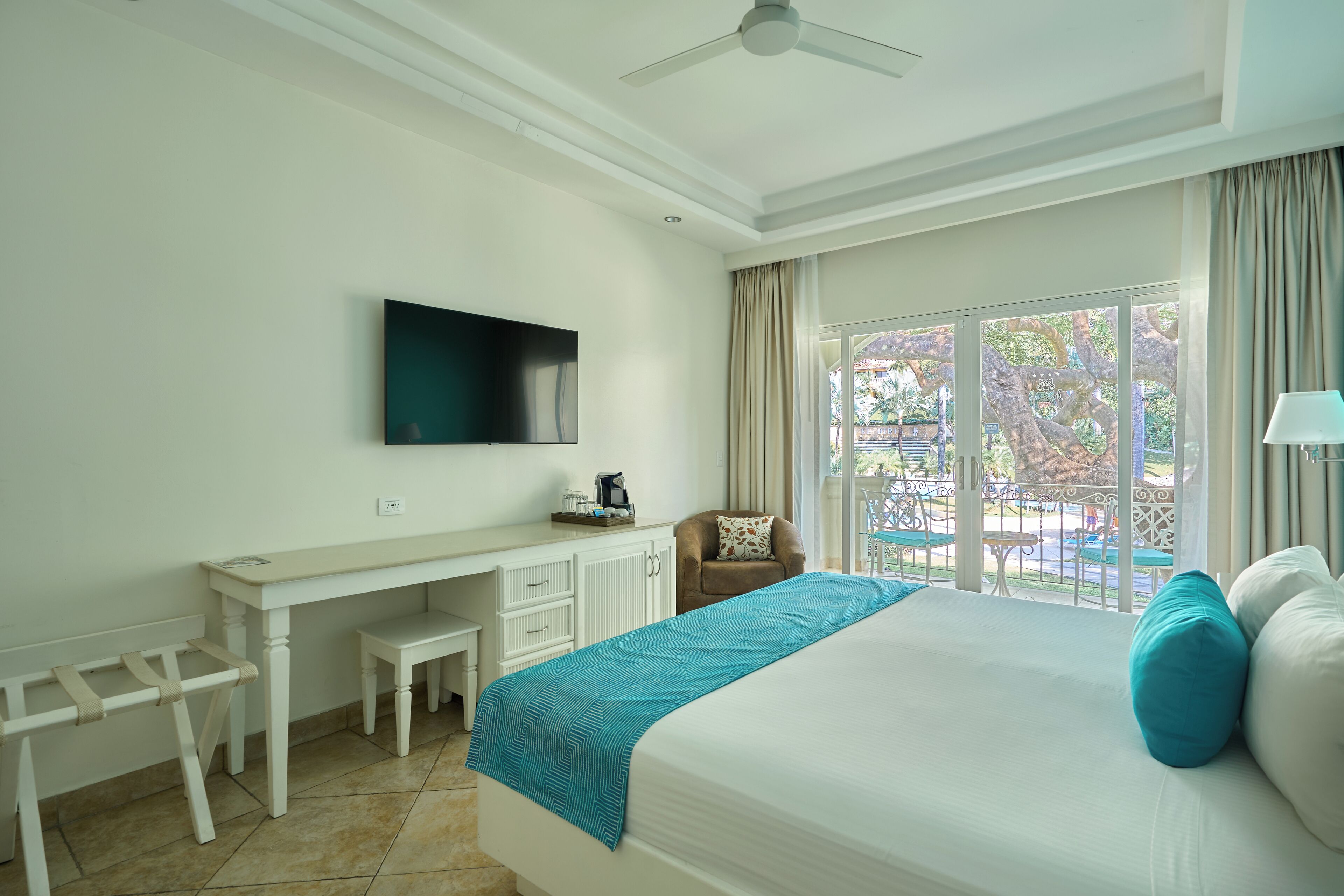 A hotel room with a bed, a desk, a chair, a TV, and a balcony with a view of trees and a pool.
