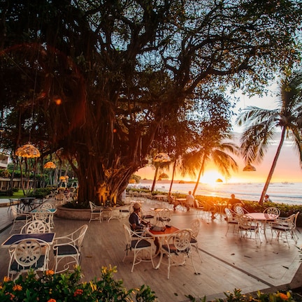 An outdoor dining area with tables and chairs, surrounded by palm trees and a view of the sunset over the ocean.