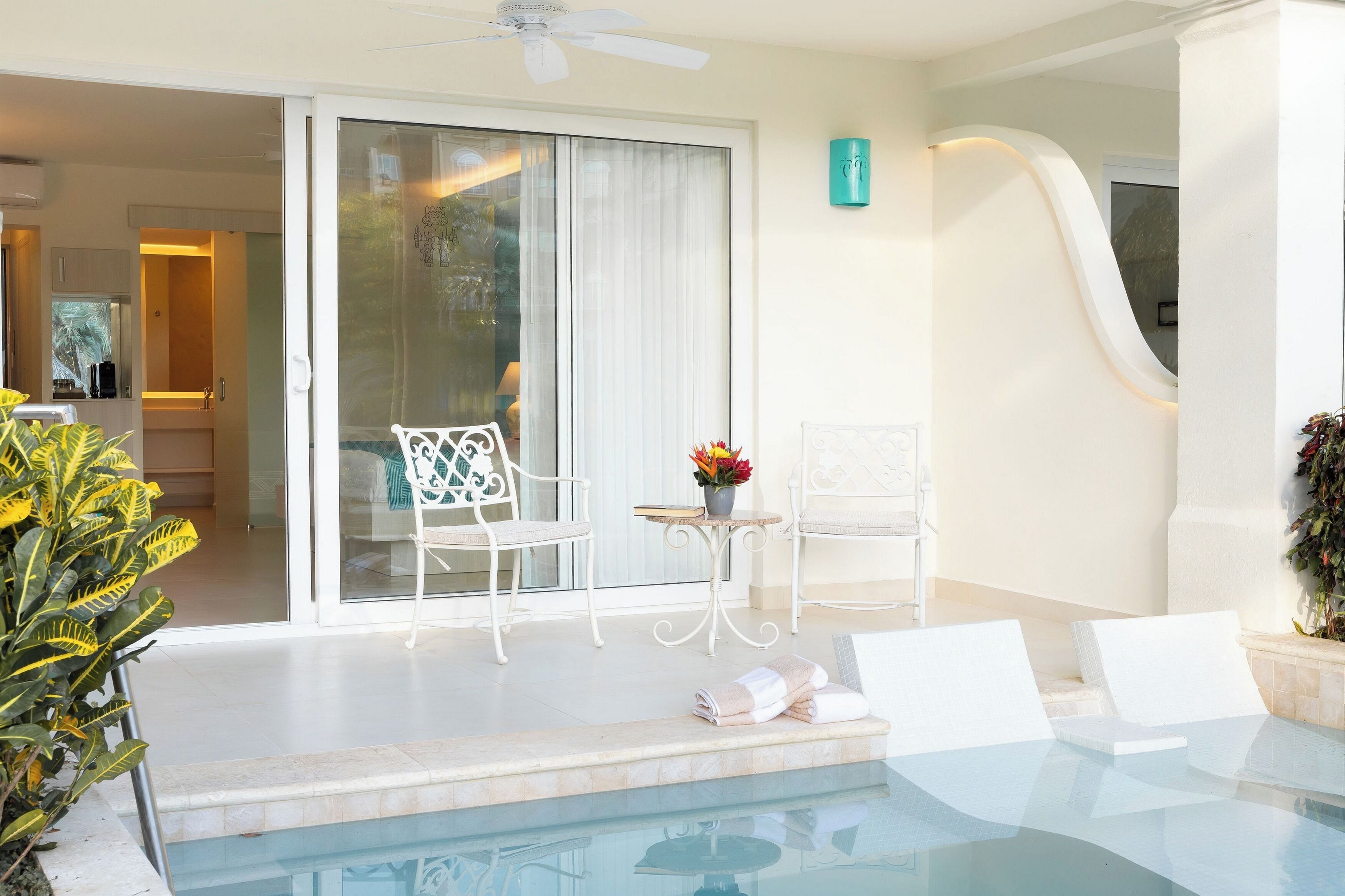 A pool area with a glass barrier, two white chairs, a small table with a vase of flowers, and a ceiling fan.