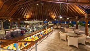 Eine Bar mit Holzbalkendecke und einer beleuchteten Bar, die in verschiedenen Farben leuchtet. Um die Bar herum stehen Korbstühle und Tische.