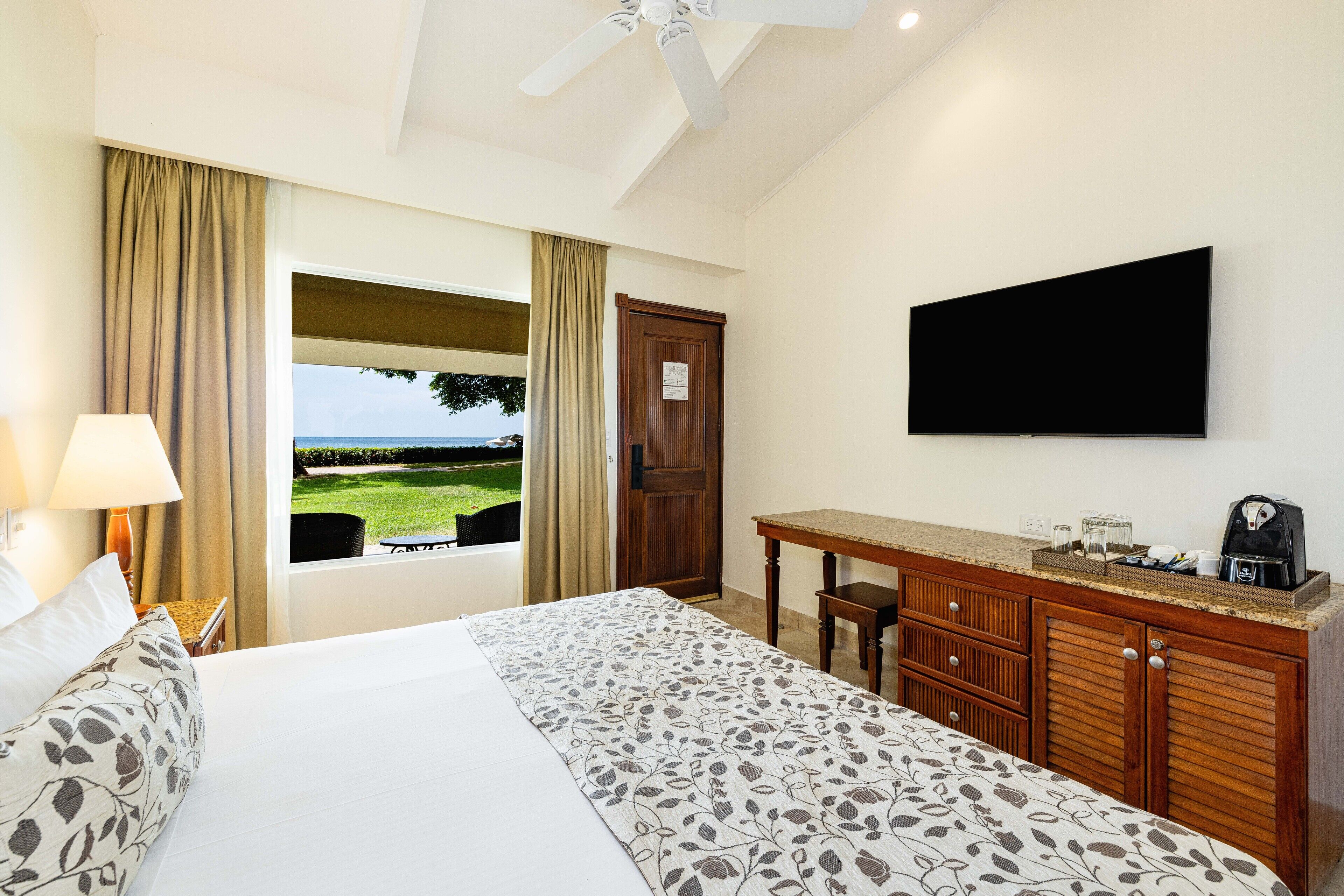Ein Hotelzimmer mit Bett, Schreibtisch mit Flachbildfernseher, Fenster mit Blick ins Freie und Deckenventilator.