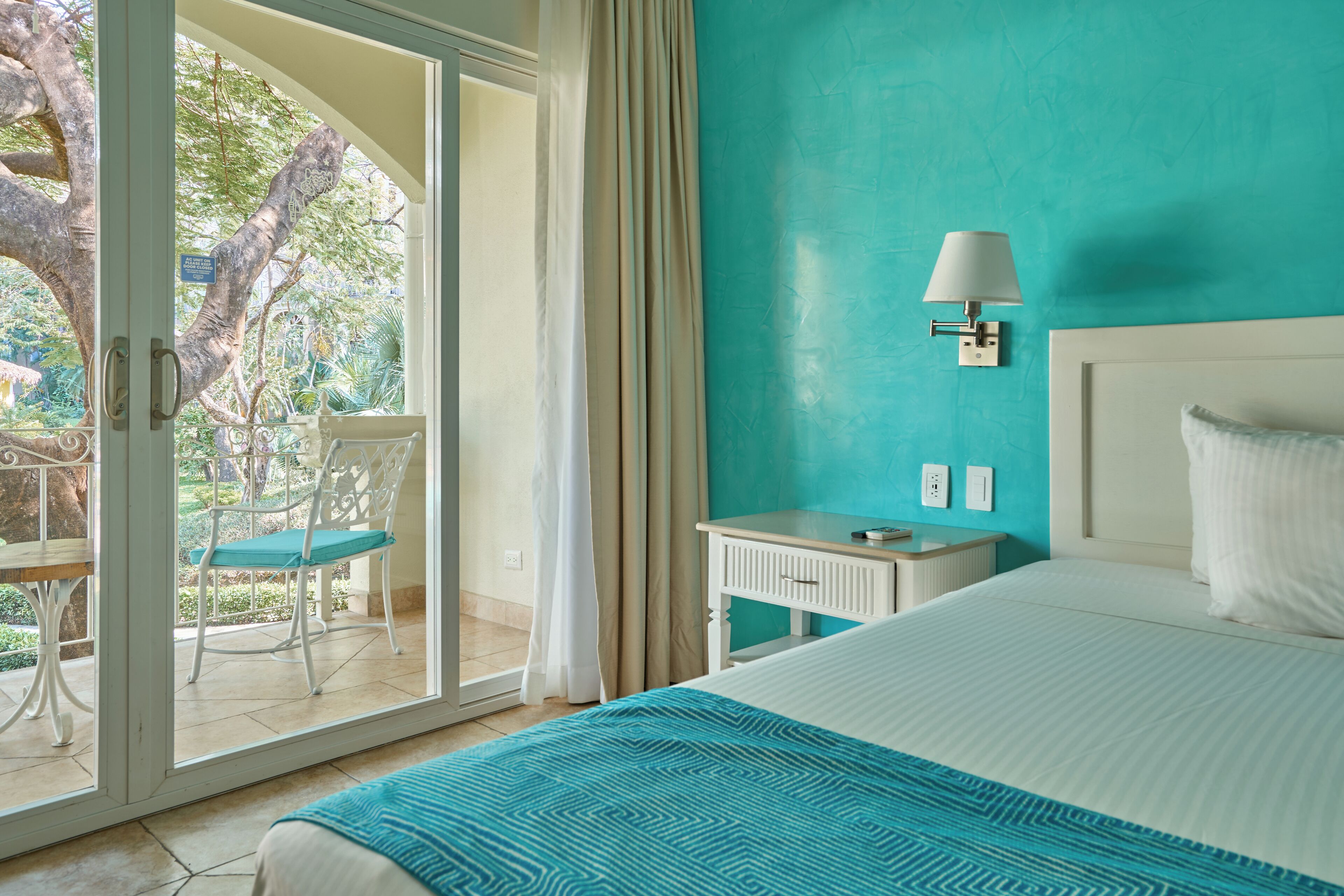 A hotel room with a bed, a nightstand, a lamp, a chair, and a view of a garden through a glass door.