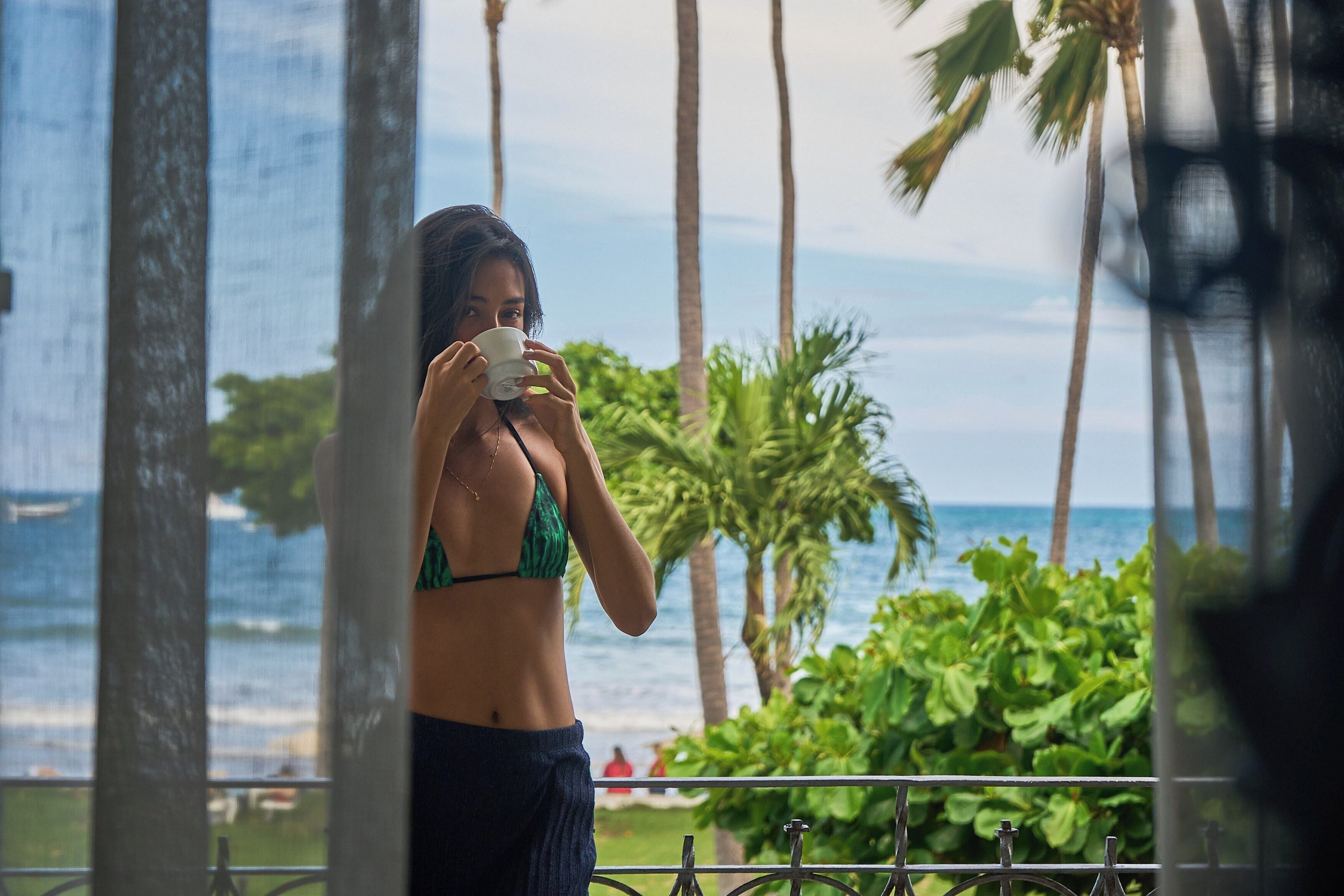 A person in a bikini standing by a window with a view of the ocean and palm trees.