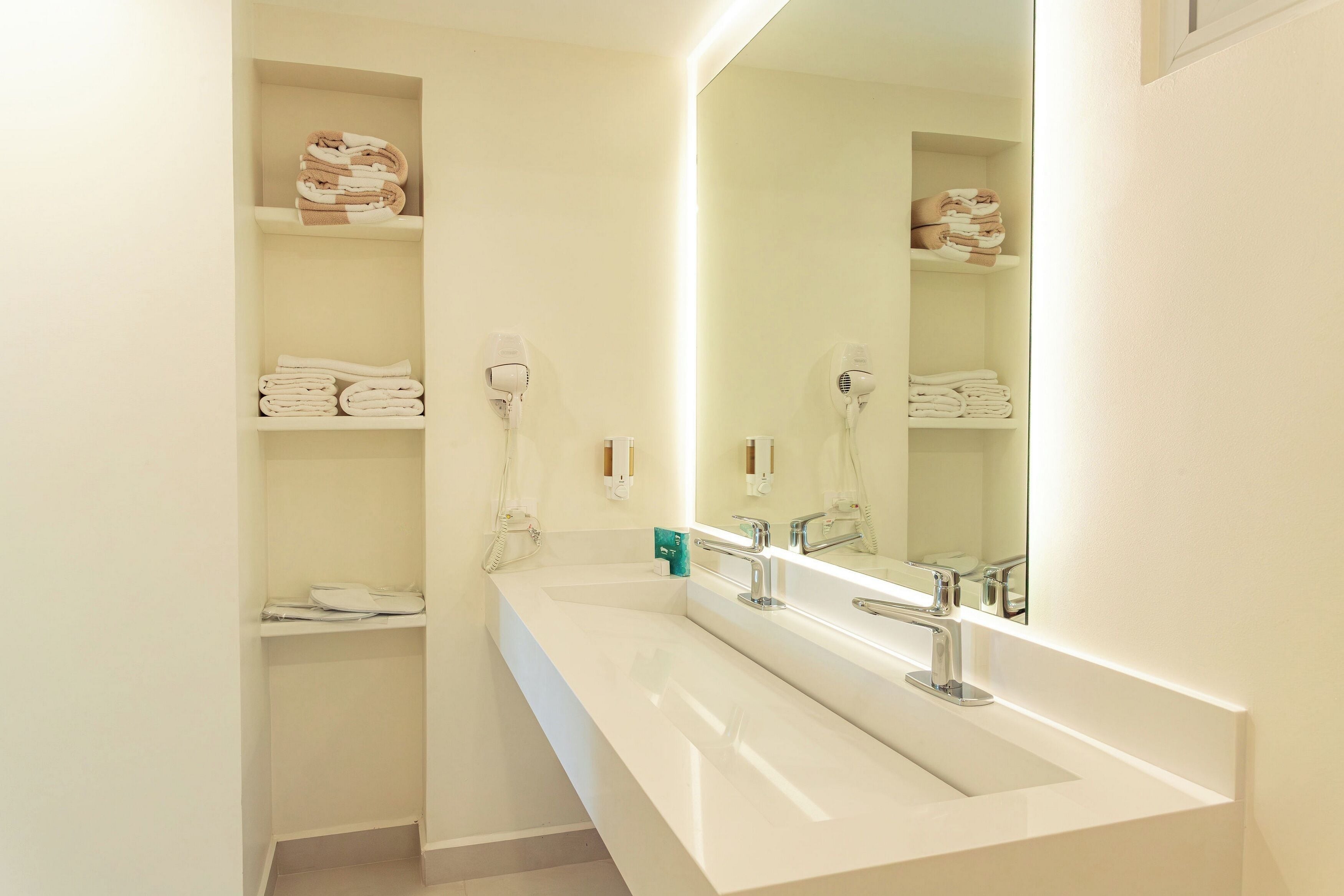 Un baño moderno con un lavamanos blanco, dos secadores de pelo y repisas con toallas dobladas.